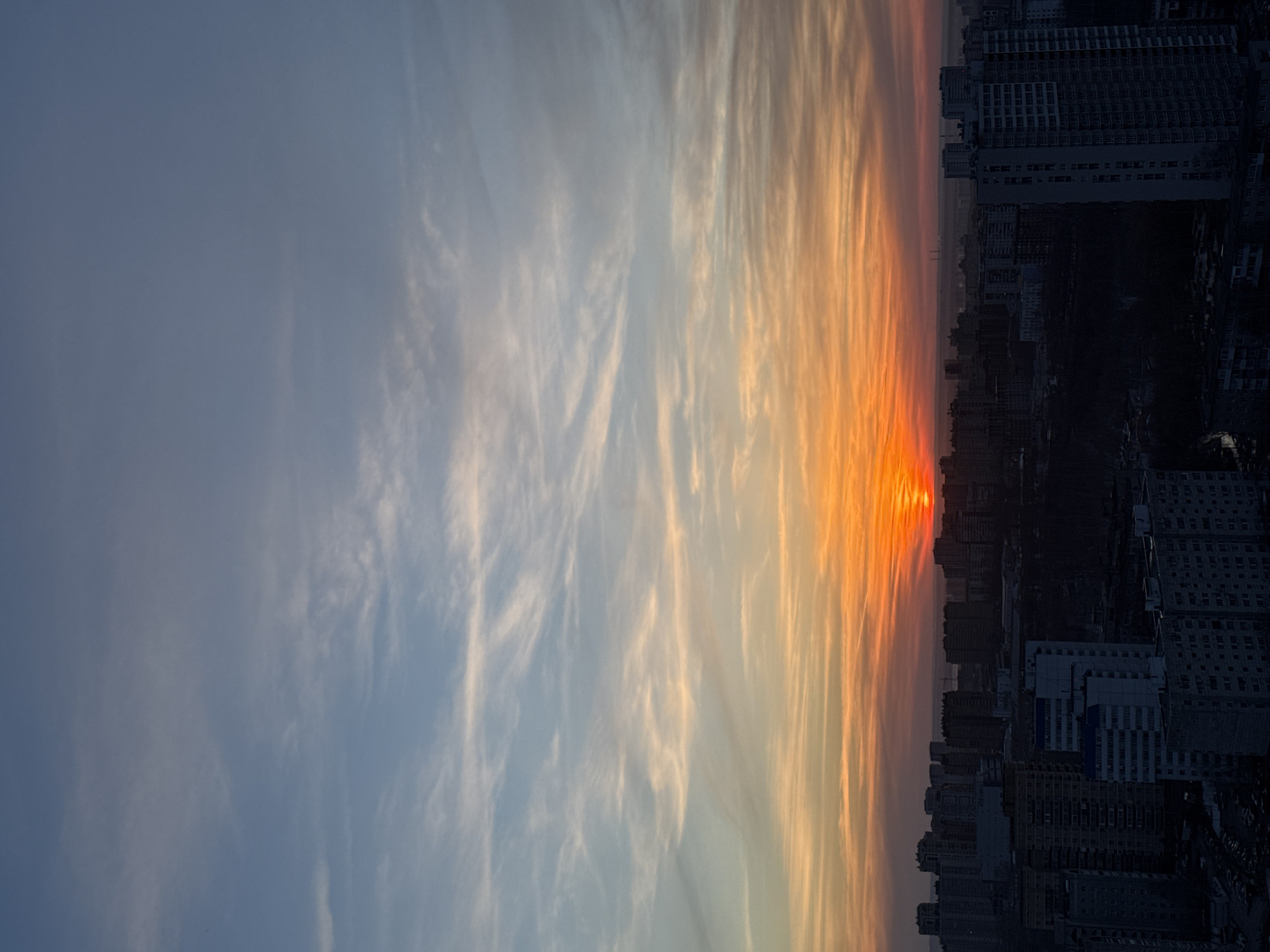 An aerial photograph captures a wide urban panorama of Perm, Russia, at either sunrise or sunset. The lower third of the image displays a dense cityscape, primarily composed of multi-story residential and commercial buildings rendered in dark silhouette against the illuminated sky. The architecture appears predominantly concrete, with visible window patterns on some structures. Clustered buildings fill the midground, extending into a hazy, indistinct background where faint industrial elements, possibly chimneys, are discernible on the distant horizon. No individuals or specific actions are visible within the urban environment. The upper two-thirds of the image depict the sky, transitioning from an intense orange and red hue at the horizon, where the sun is visible as a bright, partially obscured orb, upwards through shades of yellow and light blue, culminating in a pale blue and gray at the zenith. The sky contains scattered, wispy cirrus and altostratus clouds, elongated horizontally, which are illuminated by the low-angle sunlight, creating streaks of lighter color against the atmospheric gradient.