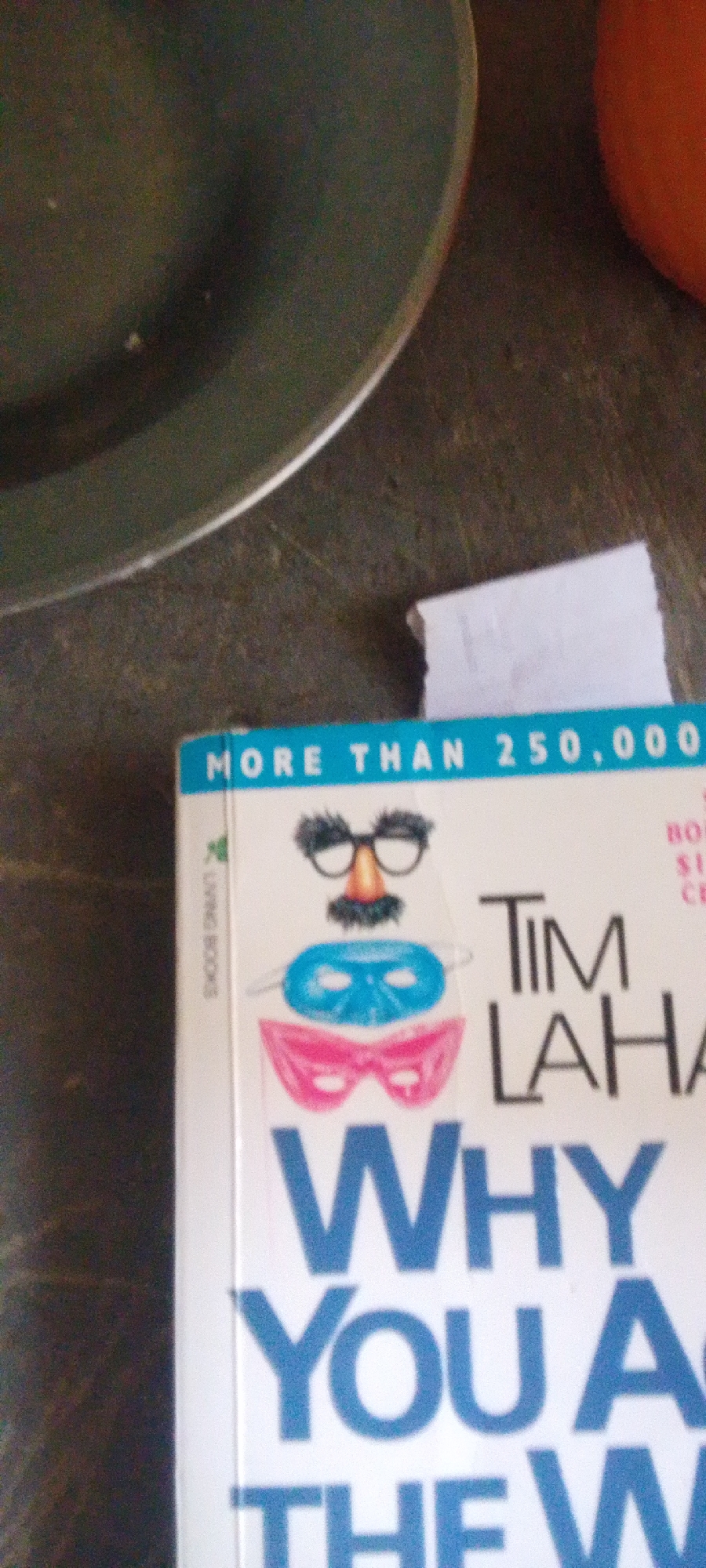 In a close-up, overhead shot, a book titled "WHY YOU ARE THE WAY YOU ARE" by Tim Lahay is partially visible. The book's cover features a collage of masks, including a Groucho Marx-style disguise, a blue mask, and a pink mask. Text on the cover reads "MORE THAN 250,000." To the left of the book, a portion of a dark green bowl or plate is visible. A sliver of a bright orange object, possibly a fruit, is also present on the right side of the frame. The background is a dark, textured surface.
