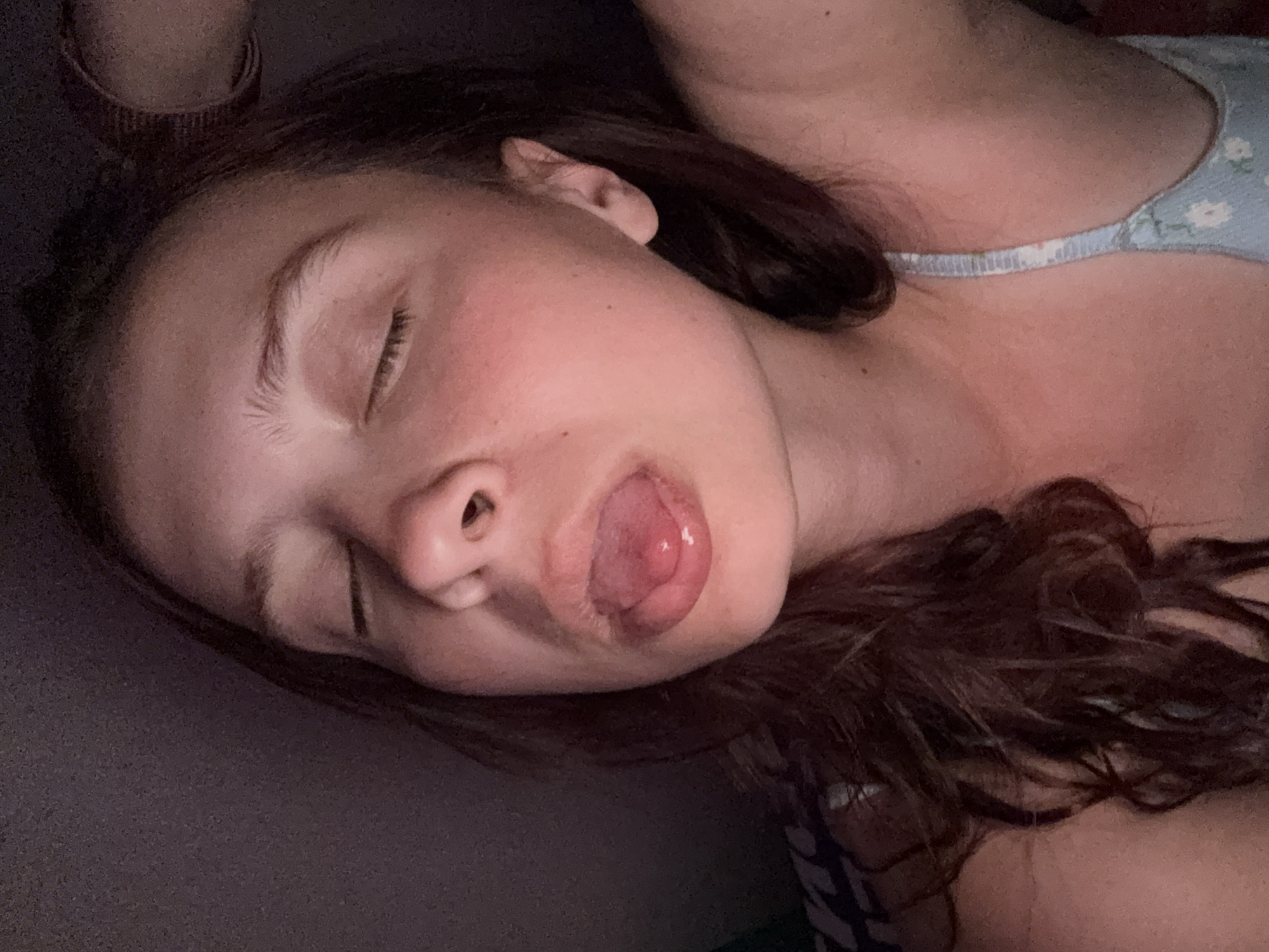 A young woman is lying down, her eyes closed, with her mouth slightly open, revealing her tongue. She is positioned at an angle, with the left side of her face closer to the viewer. Her hair is dark brown and curly. She wears a light-colored top with a floral pattern. The background is indistinct, but a dark-colored fabric or surface is visible behind her head. The location is unknown, Прикамский, Russia, is not visible in the image.