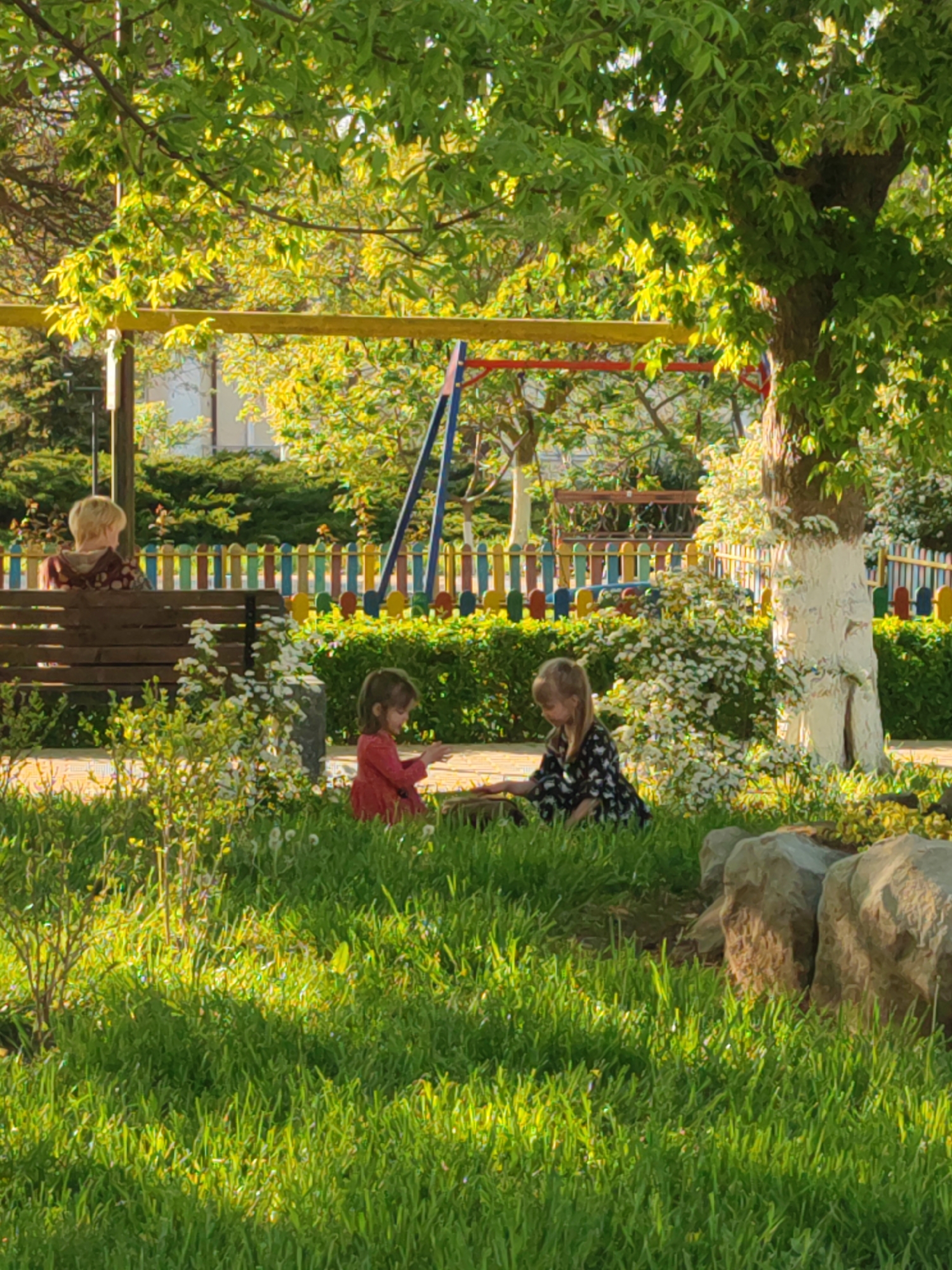 Two young girls are playing on the grass in a park. One girl is wearing a red dress, and the other is wearing a black and white patterned dress. Behind them, an adult sits on a park bench near a colorful fence. A playground with a yellow bar and blue swing set is visible in the background. The location is Sennoy, Russia.