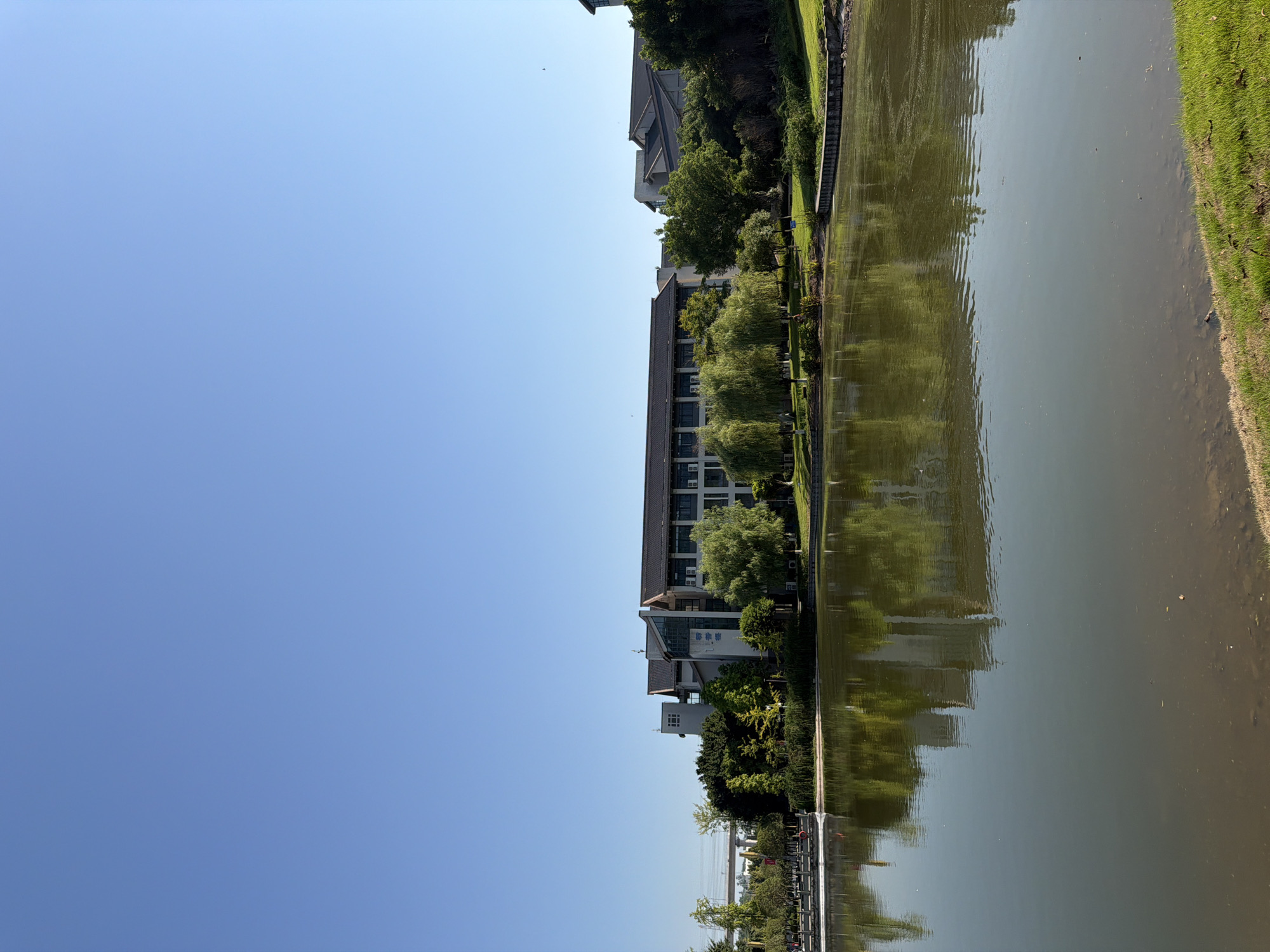 In the city of Jiangjin, China, a serene outdoor scene unfolds with a tranquil lake occupying the foreground. The lake's surface, a muted green, reflects the clear blue sky and the surrounding environment, with subtle ripples disturbing the perfect symmetry. Along the far bank, a lush green lawn slopes down to the water's edge, interspersed with patches of dry leaves and grass. Behind the lawn, a dense line of trees, predominantly willows with their characteristic drooping branches, forms a verdant backdrop.
Beyond the trees, a modern, multi-story building complex stands, characterized by its dark tiled roofs and white-walled facades. Some windows are visible, and air conditioning units are mounted on the exterior walls. The architecture appears to be institutional or educational, with clean lines and a functional design. To the left of the main building, a glimpse of a bridge or overpass can be seen, suggesting proximity to urban infrastructure. The sky above is a vast expanse of uninterrupted blue, a symbol of a clear, bright day. There are no people visible in the scene, contributing to the overall sense of calm and stillness. The image captures a moment of peaceful coexistence between nature and human construction, showcasing a well-maintained park or campus setting.