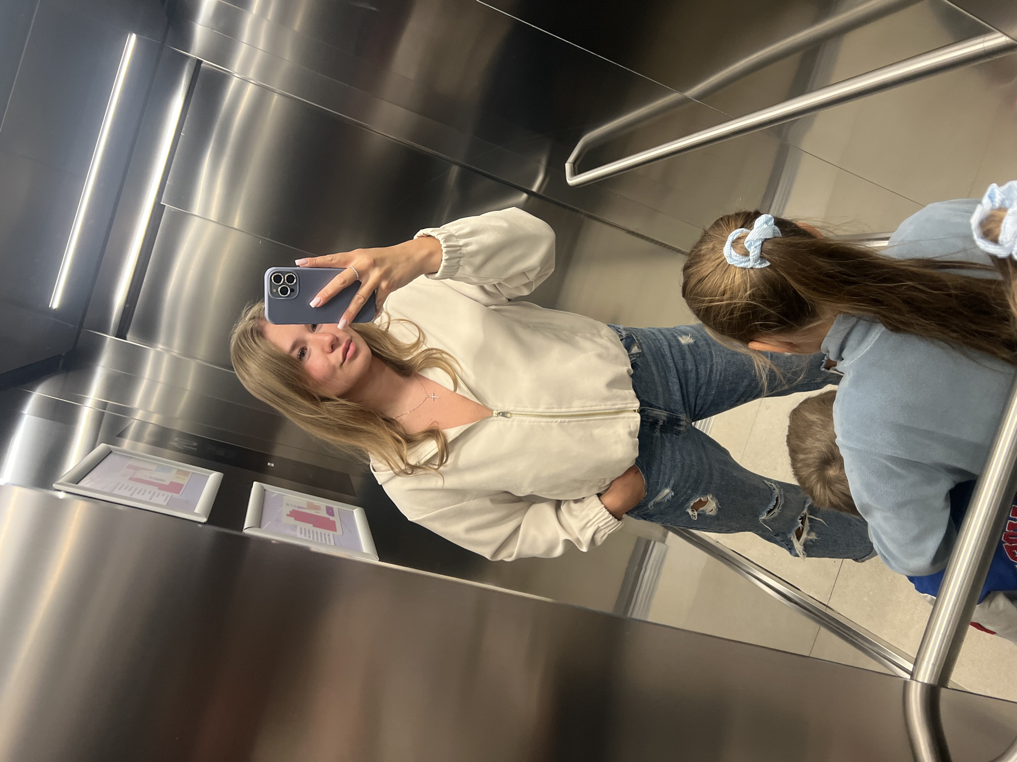 The image shows an interior space, likely an elevator, with mirrored stainless steel walls. The scene is dominated by two females and a male, possibly a family group. One female is taking a selfie with her phone, while the other female is looking at the floor, two blue hair clips on her hair. The third person, a male, is wearing blue and red clothing, sitting on the floor. There are two framed signs on the wall.
The location in the city of Budva, Montenegro cannot be identified from this image.
