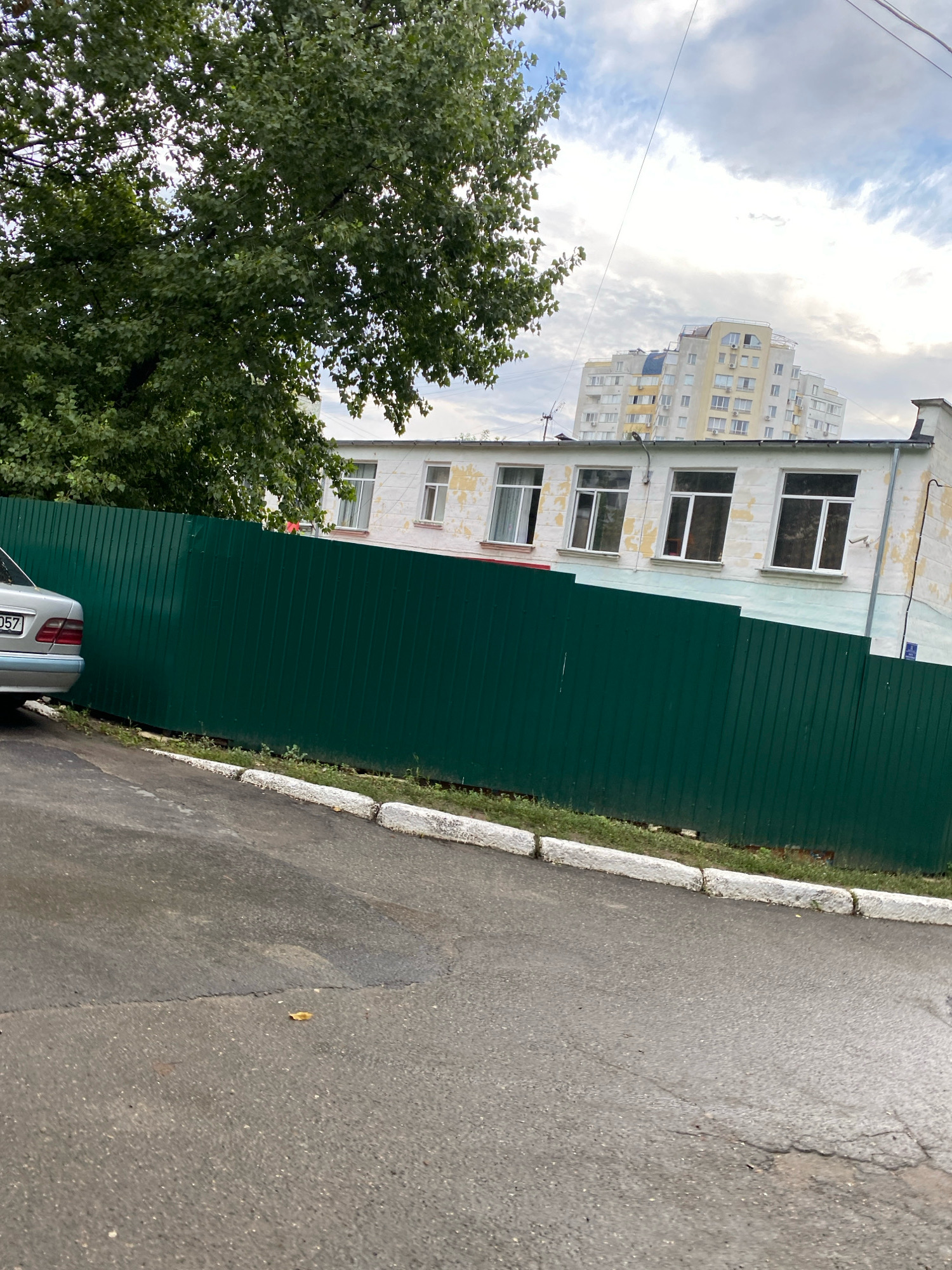 In Chișinău, Moldova, an outdoor scene captures a stretch of asphalt road with a slight incline, bordered by a low, white-painted curb. Beyond the curb, a dense, dark green metal fence runs horizontally across the frame, obscuring much of the background. To the left of the fence, the rear of a silver sedan is visible, its license plate partially obscured. Above the fence, a two-story building with weathered white walls and several windows is seen. In the distance, a modern, multi-story apartment building with a light yellow and white facade stands against a cloudy sky. A large, leafy green tree dominates the upper left portion of the image, its branches extending over the fence and partially obscuring the building. Overhead, power lines are strung across the sky.