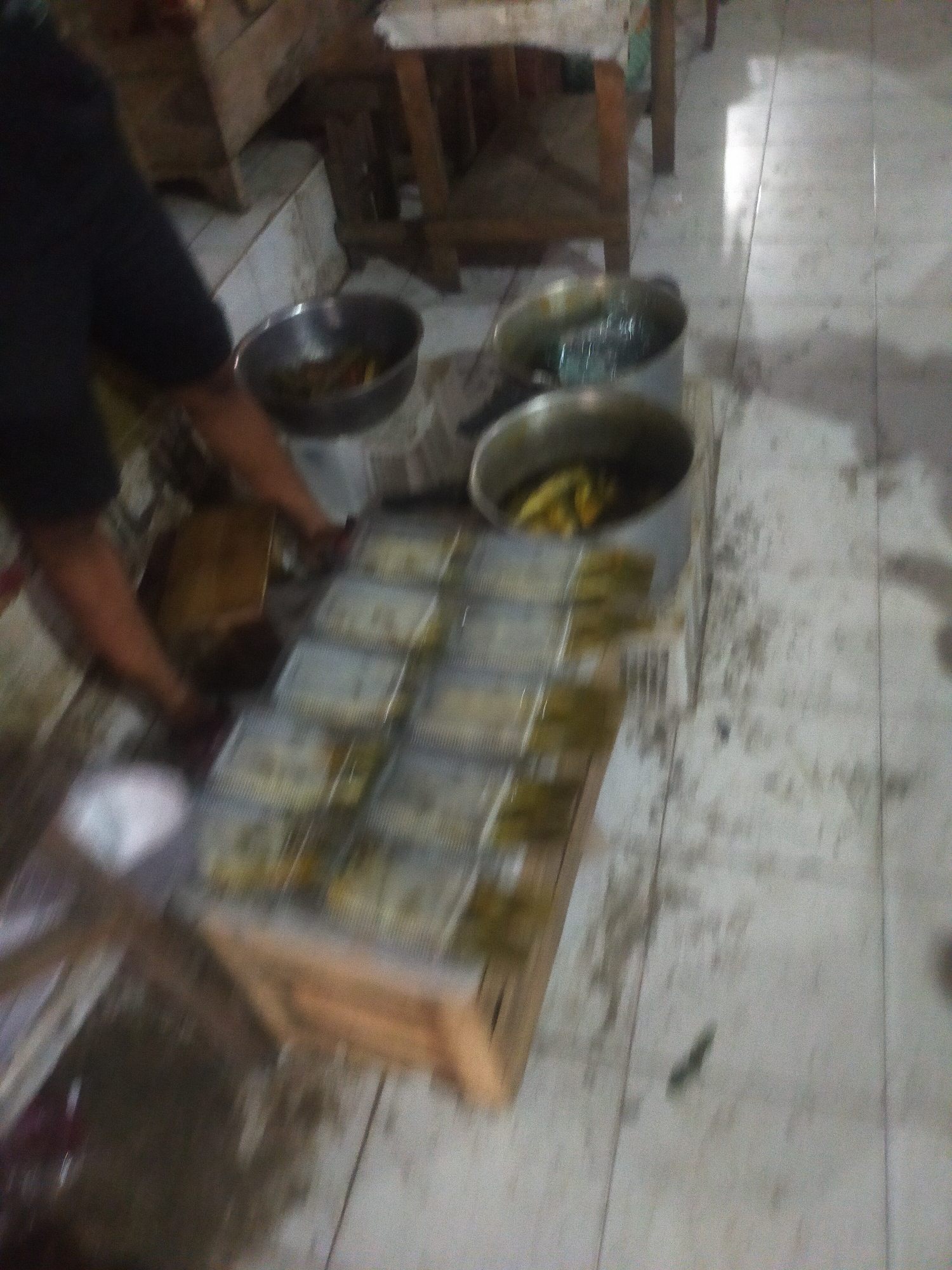 In a room with tiled flooring, a person is preparing food. The scene suggests a commercial kitchen or food preparation area. Several containers of food are arranged in rows on a wooden surface, indicating that meals are being packaged for distribution or sale. Two large pots, one containing what appears to be cooked food, are also present. The overall lighting is dim, and the image quality is poor due to blurriness, making precise identification of specific items difficult. The location is identified as Trayeman, Indonesia, though no specific landmarks are visible to confirm this.