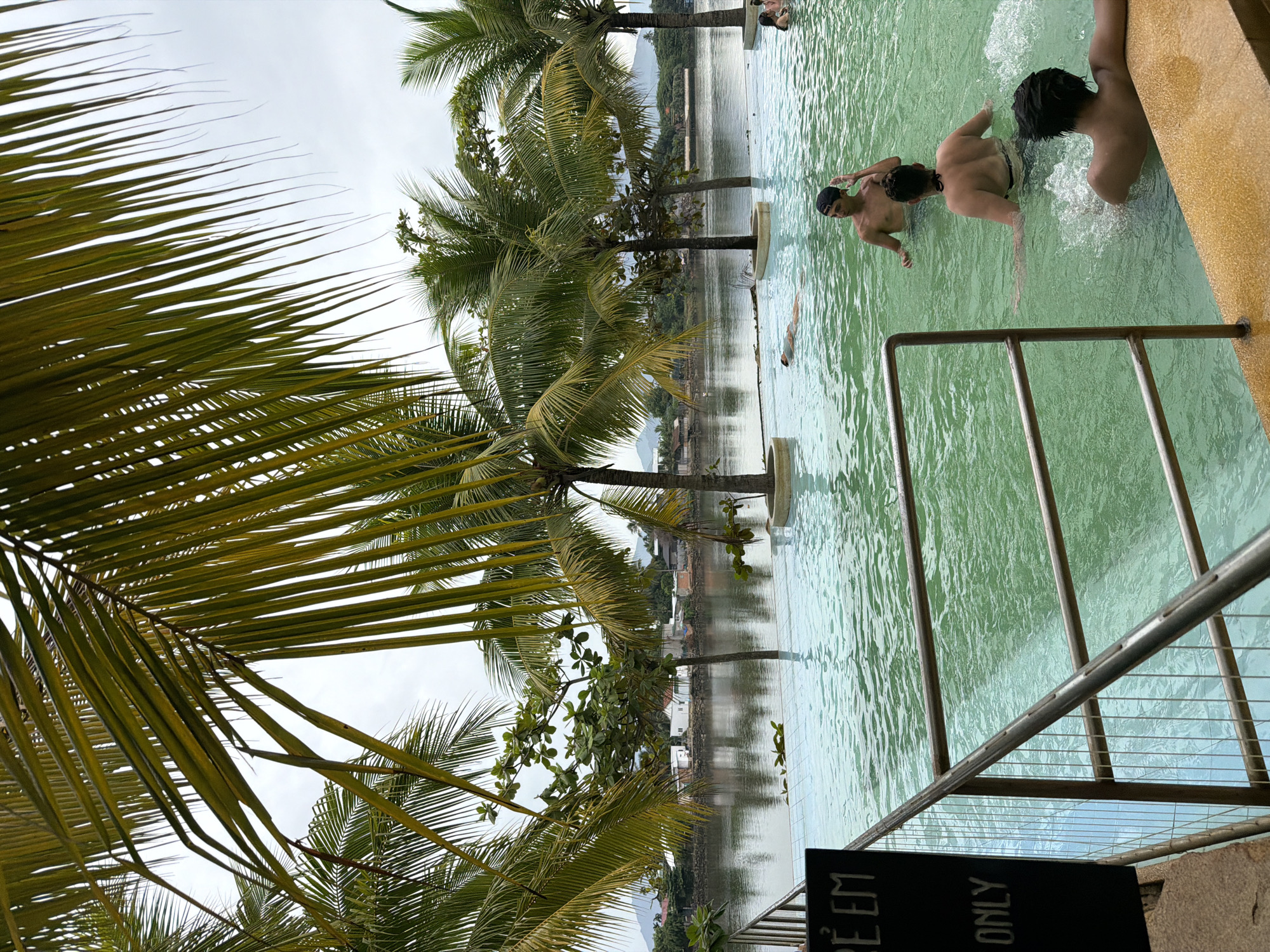 This image depicts an infinity pool overlooking a body of water in Nha Trang, Vietnam. The pool has green-tinted water and is surrounded by a tiled edge and a yellow ledge. Palm trees with green fronds frame the top of the image, and their reflections are visible in the water. In the distance, across the water, is a shoreline with buildings and lush green hills under a cloudy sky.
Several people are present in the pool. In the foreground, on the right, a person with dark, wet hair is partially submerged, leaning on the pool's edge. To their left and further back, another person in a black bikini top and dark shorts is in the water, with their back to the camera, creating ripples. A young male wearing a black baseball cap is standing in the water, facing the camera and touching his head. Further in the background, another person is floating on their back in the water. On the far right, two more individuals are visible in the pool. A dark sign with white text that reads "RE EM" and "ONLY" is visible on the left side of the frame, attached to a metal railing. The railing itself, made of silver metal, runs along the edge of the pool.