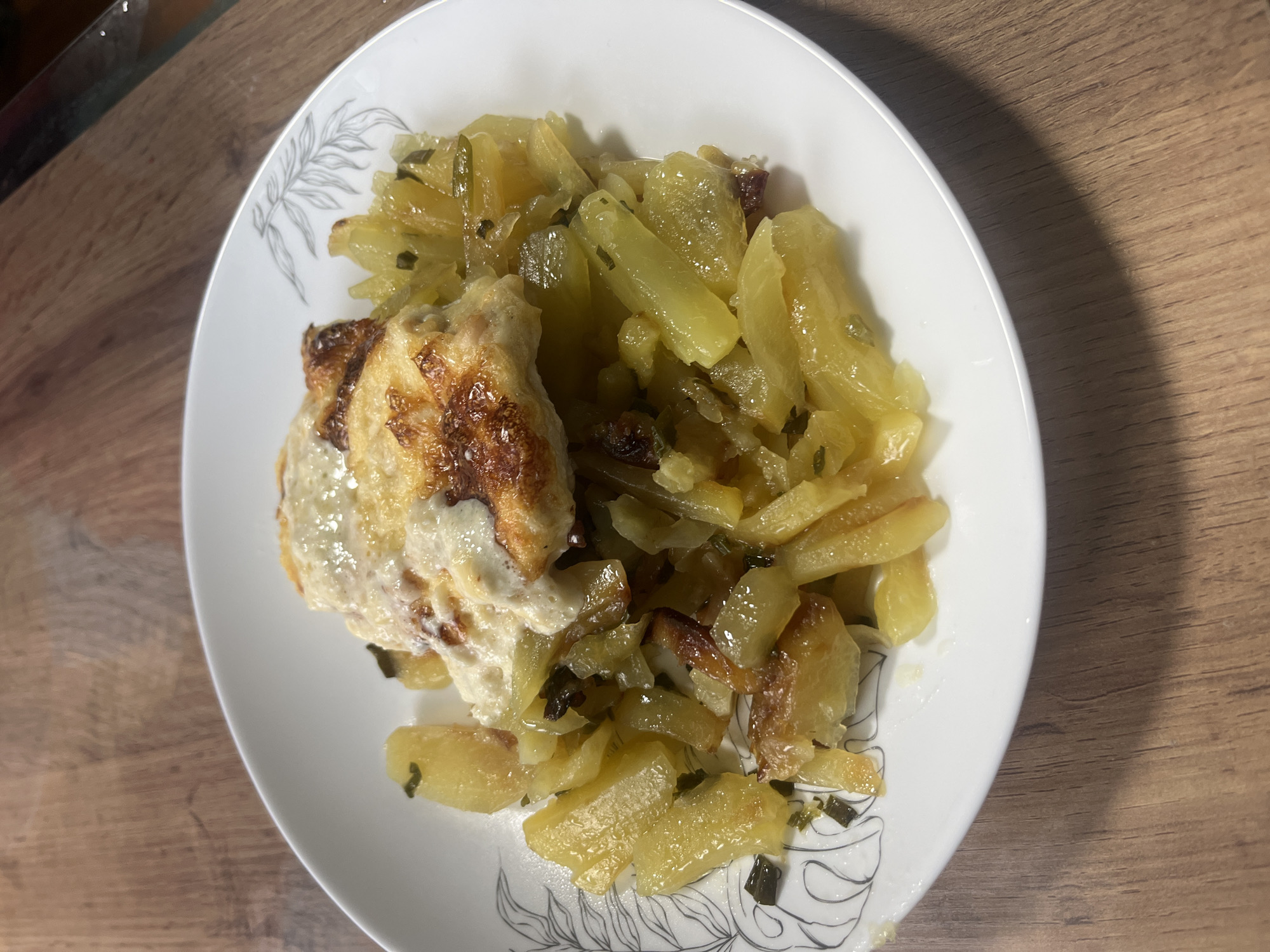 A white plate containing a serving of sliced potatoes mixed with green herbs and topped with a cheesy, baked topping rests on a wooden surface. The potatoes appear to be pan-fried or roasted, with some golden-brown edges. The topping is melted and browned, suggesting it was baked under a broiler or in an oven. The plate has a decorative rim with a subtle, etched botanical design. The wooden surface shows a grain pattern. The lighting suggests an indoor setting, possibly a kitchen or dining area. The image is a close-up shot, focusing on the food. There is no information in the image to suggest its location in Nizhny Tagil, Russia.