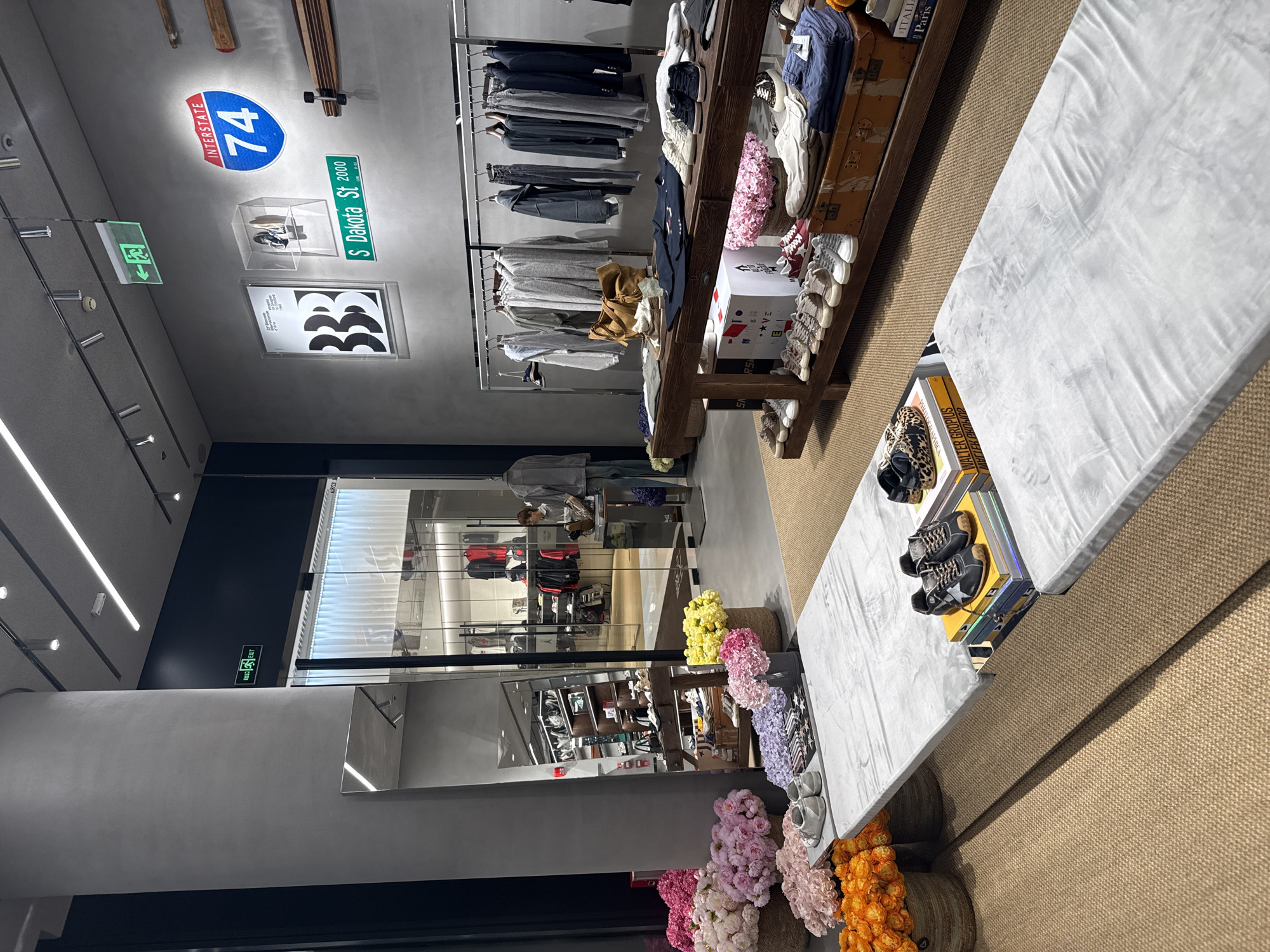 The photograph depicts the interior of a clothing store in Tianhe District, China, evidenced by the street sign "S Dakota St 2000" and the exit sign which likely indicates a commercial location. The store features clothing racks displaying various garments, shelves with shoes and accessories, and floral arrangements in baskets. A display table showcases additional merchandise. The layout suggests a retail environment designed to attract customers.