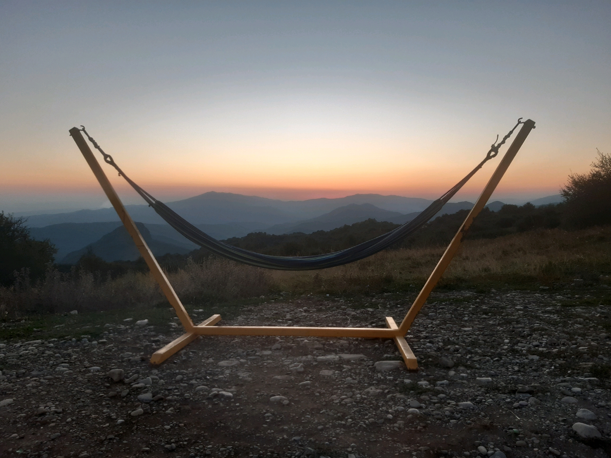 The image shows an empty hammock on a stand set up outdoors, with a scenic view of mountains and a sunset in the background. The hammock is suspended between two angled wooden supports, forming a triangular structure. The stand's base is made of wooden beams that rest on a ground covered with gravel and small stones. Beyond the hammock and stand, rolling hills and mountains are visible, fading into a soft haze. The sky displays a gradient of colors, from a pale blue at the top to a warm orange and pink band near the horizon, characteristic of a sunset. The overall setting suggests a tranquil, natural environment, possibly a campsite or a scenic overlook. There are no people, animals, or buildings present in the frame. The location is not Verona, Georgia; it is a mountainous region during sunset.