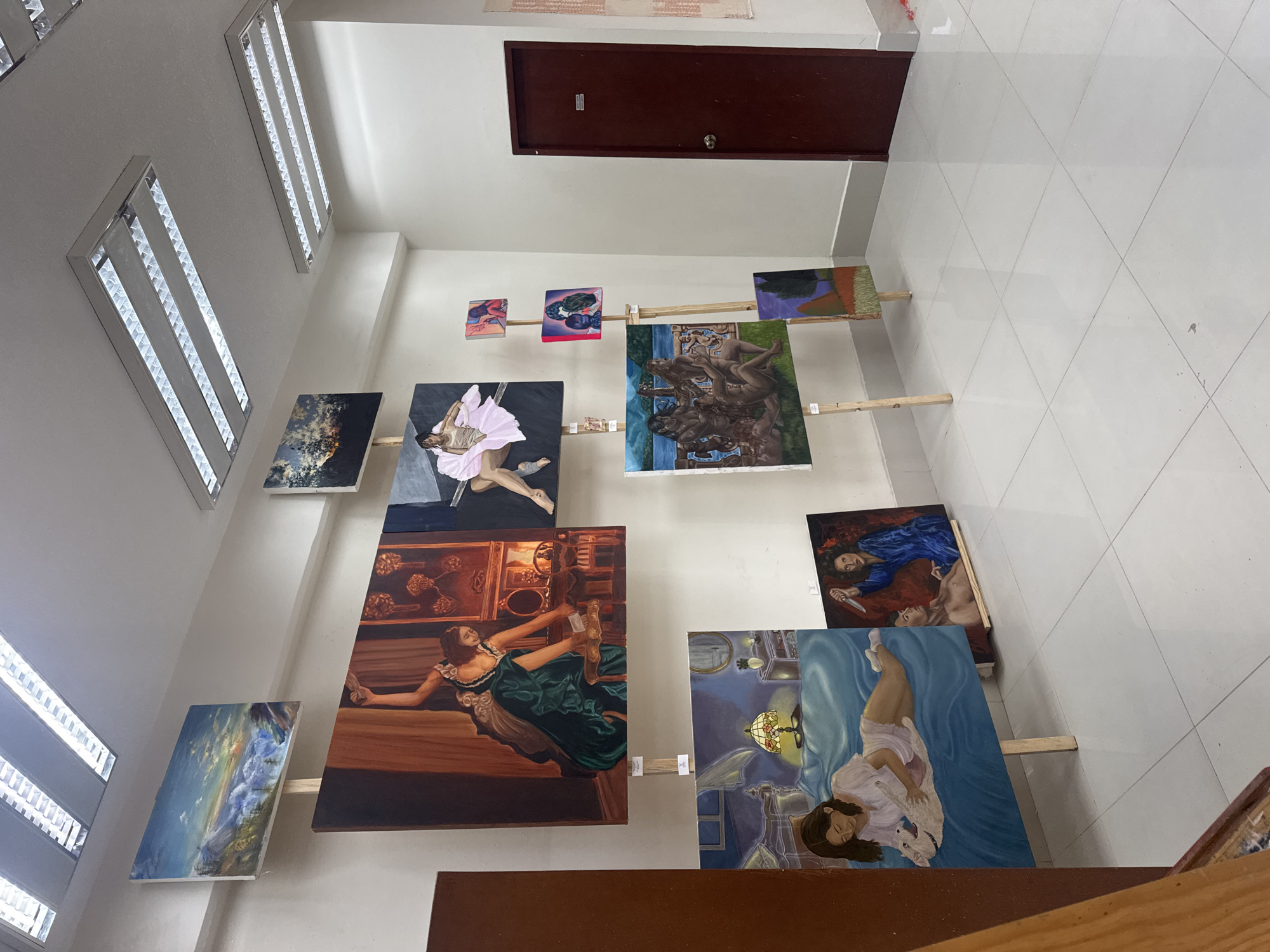 The image shows an interior view of a building in San Andrés Cholula, Mexico, featuring a series of paintings displayed on the walls. The paintings are exhibited at varying heights and appear to be supported by wooden beams. The artwork depicts a range of subjects, including figures, landscapes, and abstract compositions. The building's architecture includes fluorescent lighting fixtures and a dark-colored door. The floor is tiled in a grid pattern.