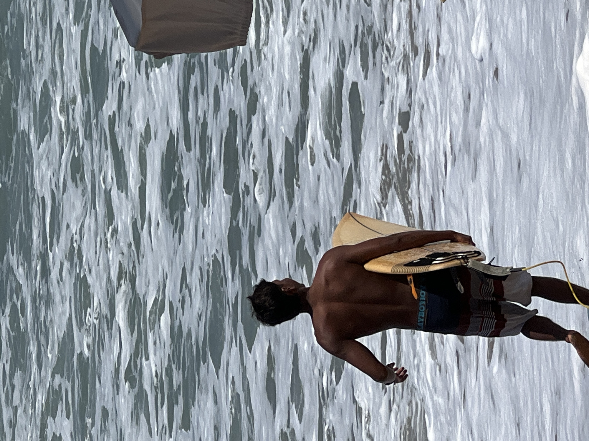 A shirtless person with dark hair is walking towards the ocean, carrying a surfboard under their right arm. The person is wearing striped swim trunks. The ocean is rough with white foam. A corner of a light-colored umbrella is visible in the upper right quadrant of the image. The scene appears to be a beach in Kuta, Indonesia, given the ocean conditions and surf culture commonly associated with the location.