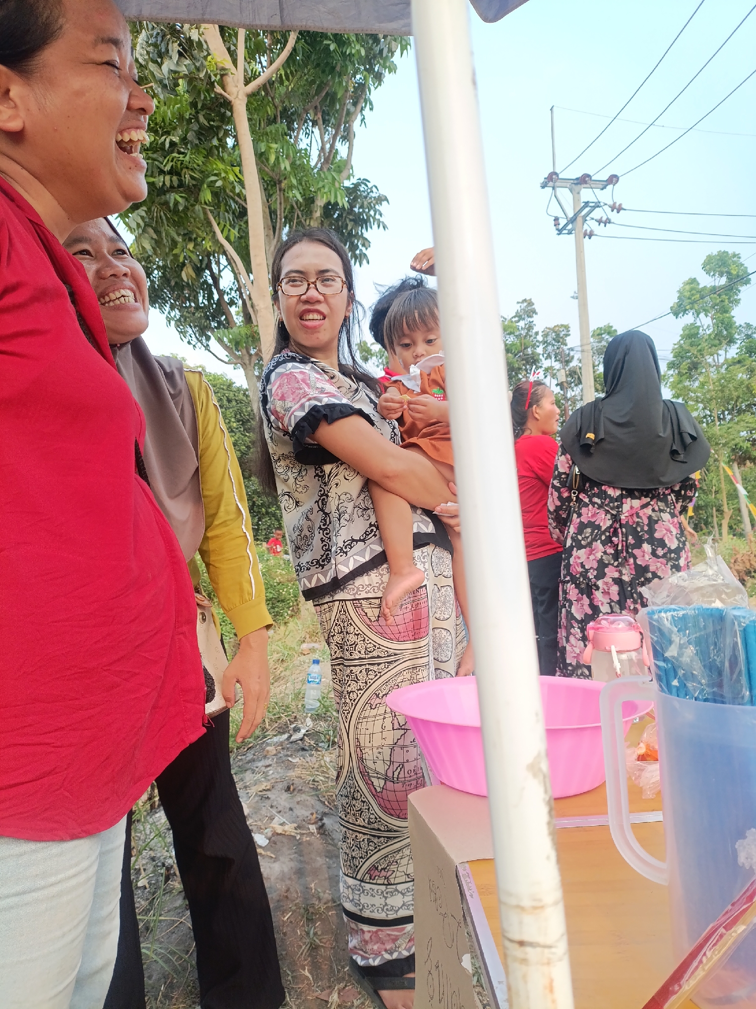 In Cariu, Indonesia, a group of people are gathered outdoors under a temporary canopy. The focus is on several women and a child. In the foreground, a woman in a bright red top laughs heartily, with another woman in a grey hijab beside her, also smiling. To the right, a woman wearing glasses and a patterned top holds a young child dressed in orange. Behind them, a woman in a black hijab and a floral dress stands next to a person in a red shirt and dark pants. The background shows trees, electrical wires, and a utility pole. On a wooden table, a pink basin and a blue pitcher filled with blue straws are visible. A bottle of water is also present on the ground.