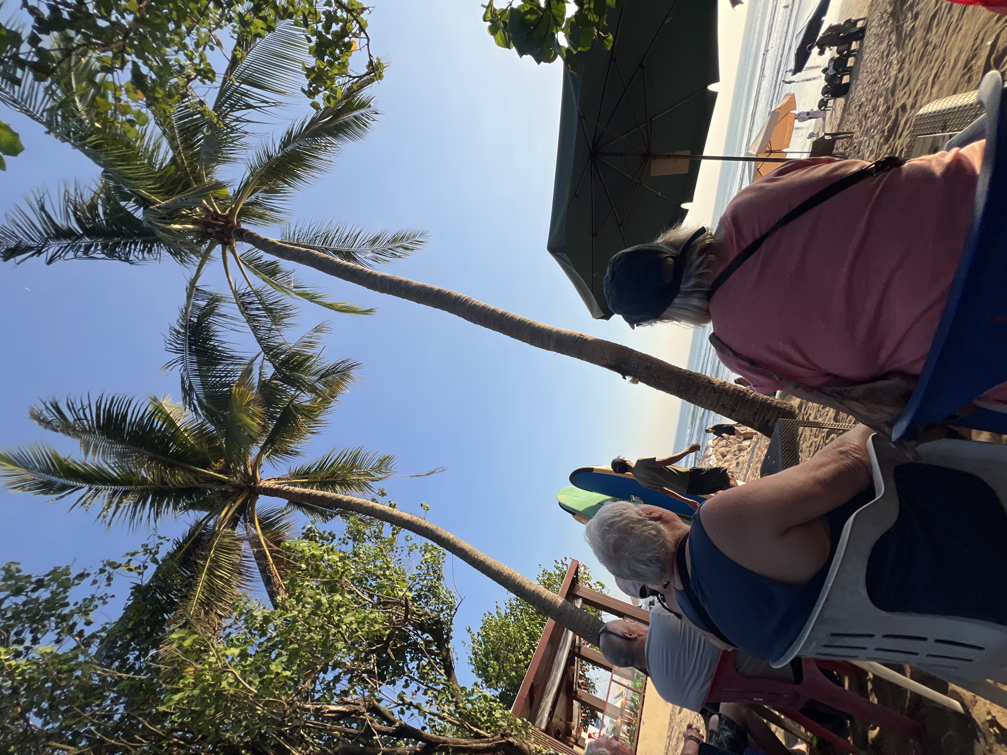 This is a photograph taken in Kuta, Indonesia, likely during sunset, depicting a beach scene with several people. In the foreground, two older individuals are seated on chairs, facing away from the camera and towards the ocean. One is wearing a blue tank top, and the other a pink shirt and a black cap. Behind them, two surfboards, one green and one blue, are propped upright. A person wearing a green t-shirt and dark shorts is walking away from the camera, seemingly towards the water. To the left, a beach hut with a red roof is partially visible, with more people seated at tables. Palm trees are prominent throughout the scene, their fronds casting shadows on the sand. In the background, the ocean stretches towards the horizon, with several small figures visible in the water. An open green umbrella shades a seating area on the beach, and a few orange umbrellas are also visible further down the beach. The sky is clear and blue, with the sun low on the horizon, indicating either sunrise or sunset.