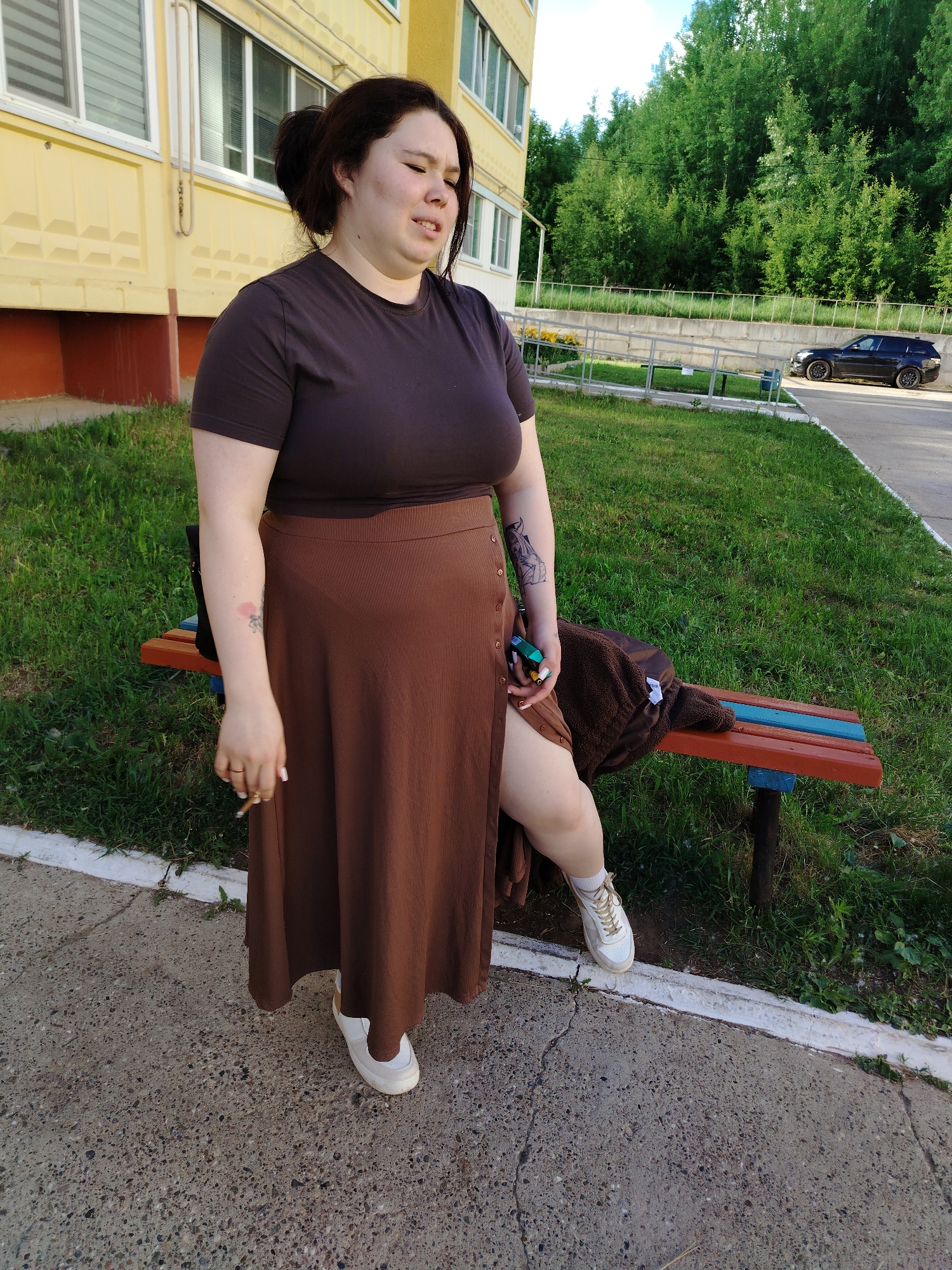 A woman with dark hair tied back stands outside in Noviy, Russia, next to a low bench. She is wearing a brown t-shirt, a brown midi skirt with buttons, and white sneakers. She is holding a lit cigarette in one hand. The bench has red and blue painted wood and a brown jacket resting on it. Behind her is a green grassy area and a beige building with windows. In the background, there are trees and a dark-colored car is visible. The ground is paved.