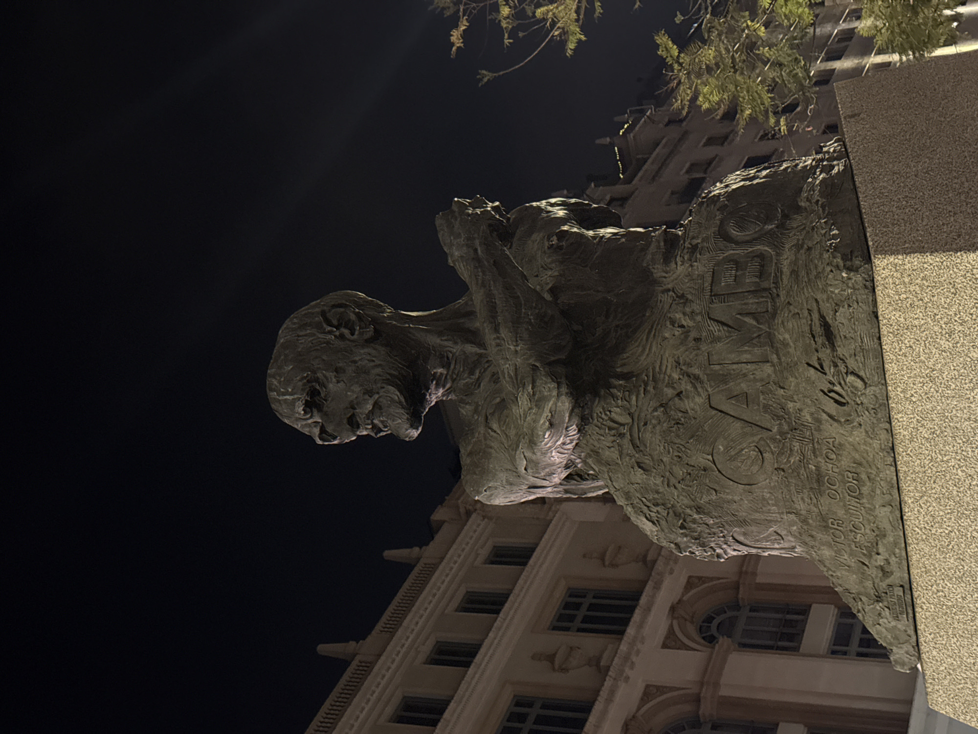 This image depicts a statue of the Chilean poet Pablo Neruda, located in Barcelona, Spain. The statue is a dark, bronze-colored sculpture of Neruda in a contemplative pose, with his hands near his face. The base of the statue bears his name, and the name of the sculptor Victor Ochoa. The statue is set against the backdrop of a light-colored building with multiple windows and architectural details, suggesting an urban environment. Above the building and behind the statue are some trees and dark night sky.