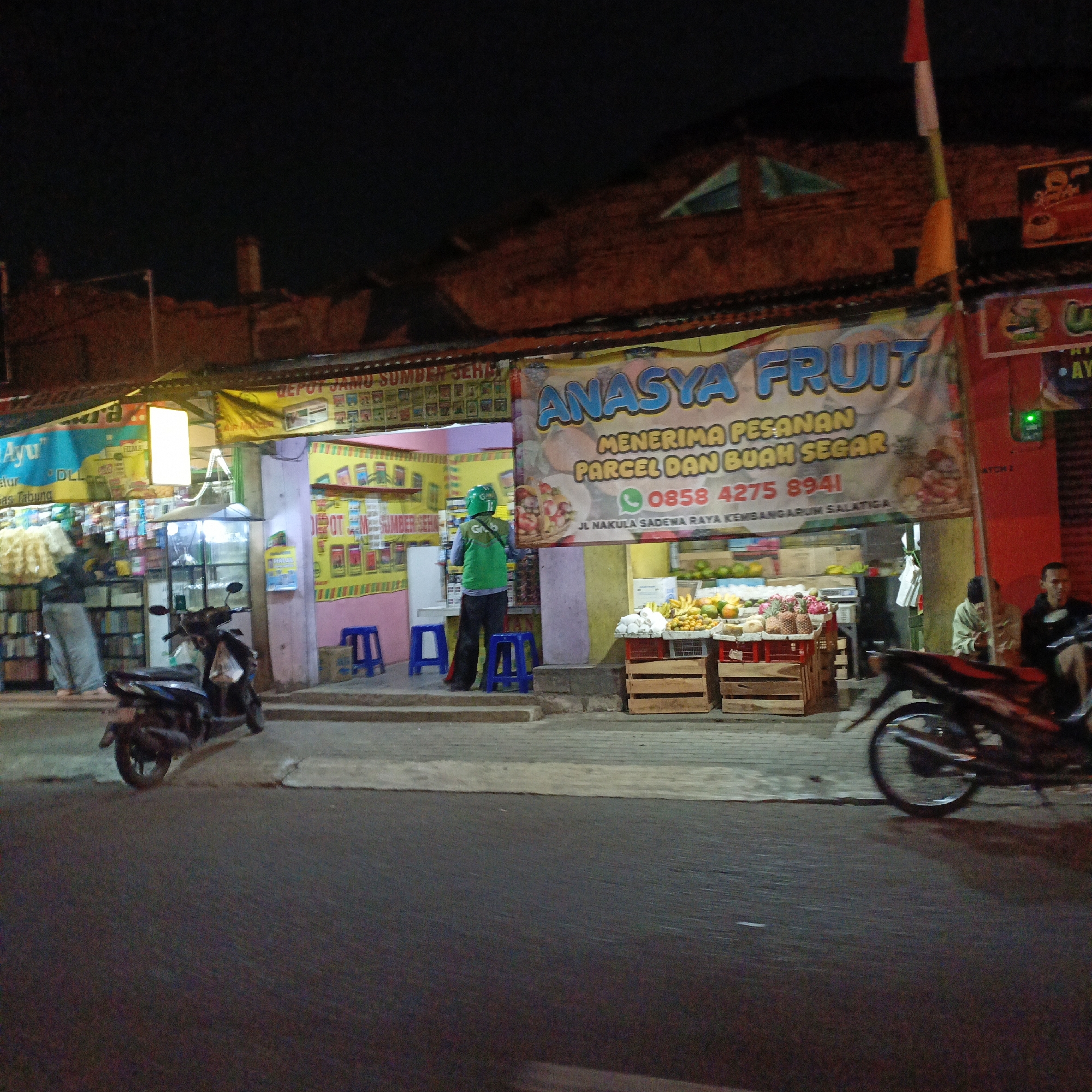 The image captures a nighttime street scene in Salatiga, Indonesia, with a focus on a fruit stall and a small shop. The fruit stall, named "ANASYA FRUIT," displays a variety of fresh produce, including bananas and pineapples, arranged in wooden crates. Above the stall, a banner advertises fruit delivery services and provides a phone number. To the left, a shop displays shelves stocked with various products. A Grab delivery driver in uniform stands near the entrance of the shop. In the foreground, two motorcycles are parked. The street is paved, and the lighting suggests it is a commercial area. The buildings in the background appear to be low-rise structures, illuminated by artificial lights. The overall atmosphere is that of a bustling local market at night.