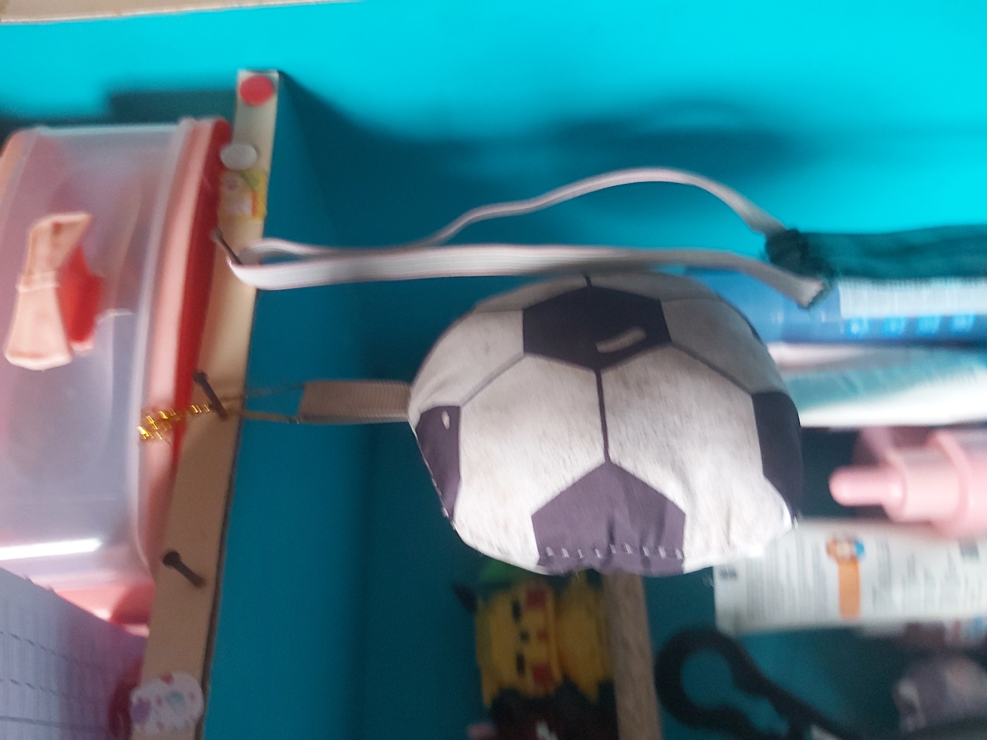 A soft, soccer ball-shaped plush toy is hanging from a shelf in a dimly lit room. The toy is white with dark purple pentagonal and hexagonal panels. A light-colored strap is attached to the top of the toy, and it is looped over a nail or hook on the wooden shelf. The shelf is light brown and appears to be a part of a larger shelving unit. Below the shelf, there are various objects, including a yellow Pikachu plush toy, a pair of black scissors, and several bottles and containers, one of which is a pink dropper bottle. The background is a bright turquoise wall. The image is slightly blurry. The location is the city of Ploso, Indonesia.