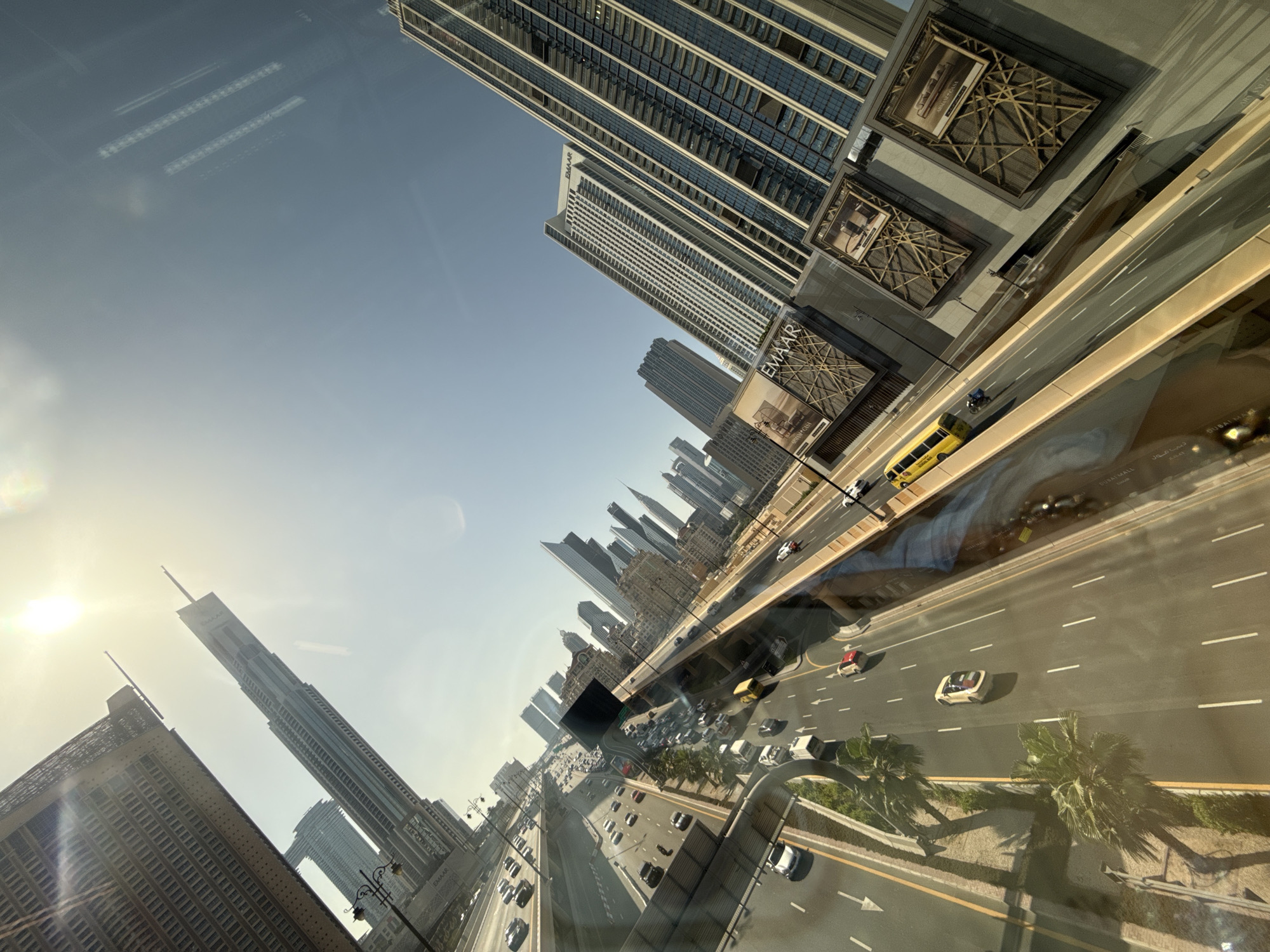 This is a high-angle, wide shot of a multi-lane highway in Dubai, United Arab Emirates, with tall modern skyscrapers lining the sides. The highway is congested with traffic, including cars, trucks, and a yellow bus. Palm trees are planted along the median of the highway. Several of the buildings display "EMAAR" branding, indicating a connection to the Emaar Properties development company. The sun is shining brightly, casting long shadows. The image appears to be taken from a window, as evidenced by reflections on the glass.