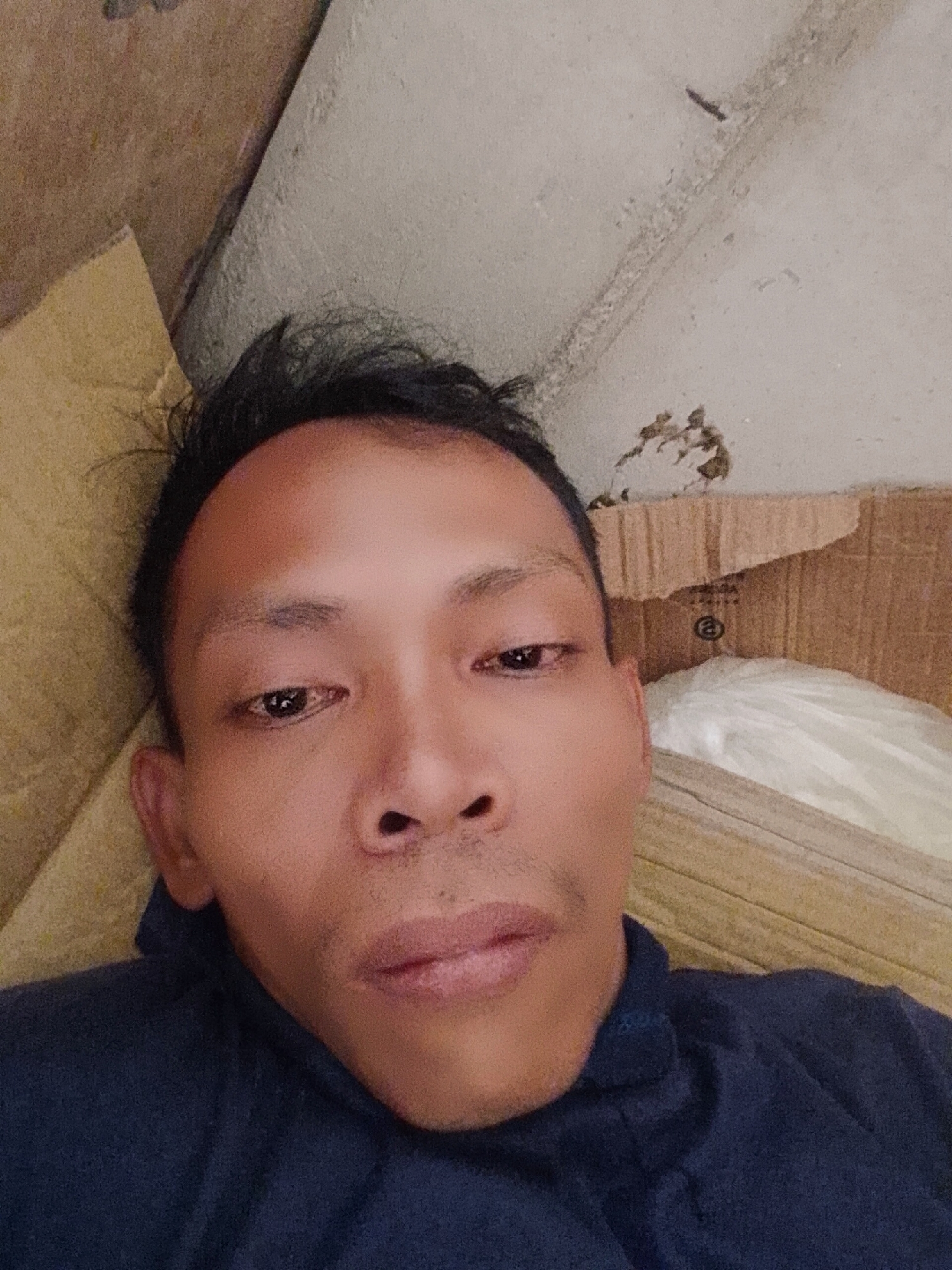 A close-up selfie image shows a man with dark hair and brown skin. He is lying down, possibly on a yellow cushion or pillow. He wears a navy blue shirt or jacket. In the background, there is a damaged wall and a piece of cardboard. It is not possible to confirm the location as Bukit Ketok, Indonesia based solely on the visual content of the image.