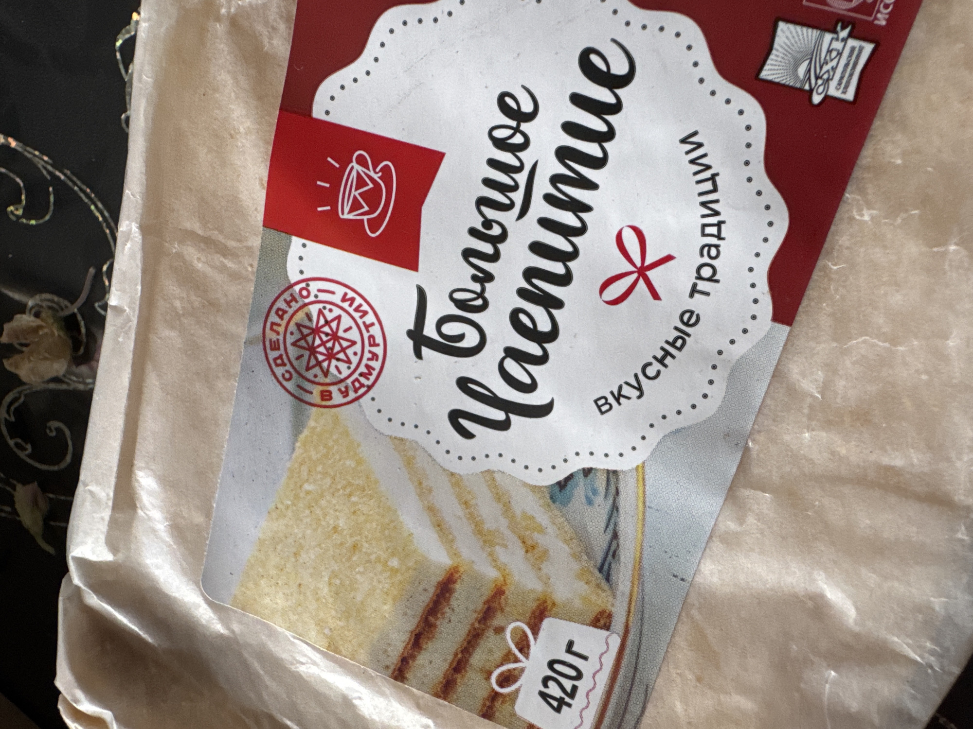 The image shows a close-up of a packaged cake. The packaging is predominantly white and red, with a picture of a slice of cake on the left side. The cake appears to have multiple layers of sponge and cream. Text in Russian is visible on the packaging, including "Большое чаепитие" (Big Tea Party), "сделано в Удмуртии" (made in Udmurtia), and "вкусные традиции" (tasty traditions). A weight indicator shows "420 г" (420g). The packaging is placed on a translucent, crinkled material, likely paper or plastic wrap, which in turn rests on a dark, patterned surface. There is no indication of the location being Старое Михайловское, Russia, or any people or actions occurring.