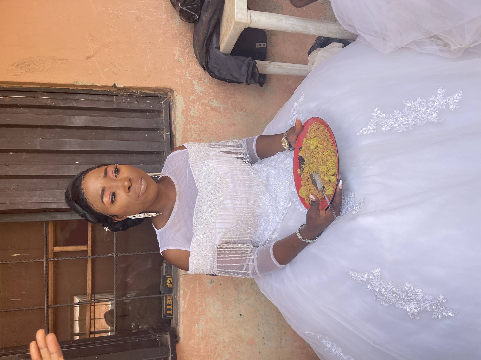 A bride in a white wedding dress sits outdoors on what appears to be a chair or bench in Akwanga, Nigeria, near a window with dark horizontal bars and an adjacent tan-colored wall. She is holding a red plate filled with a yellow and dark food item, and she is using a spoon to eat. Her hair is styled up and back, and she is wearing large, dangling earrings and a silver bracelet on her left wrist. The dress has intricate white embellishments on the bodice and skirt, and the sleeves are sheer.