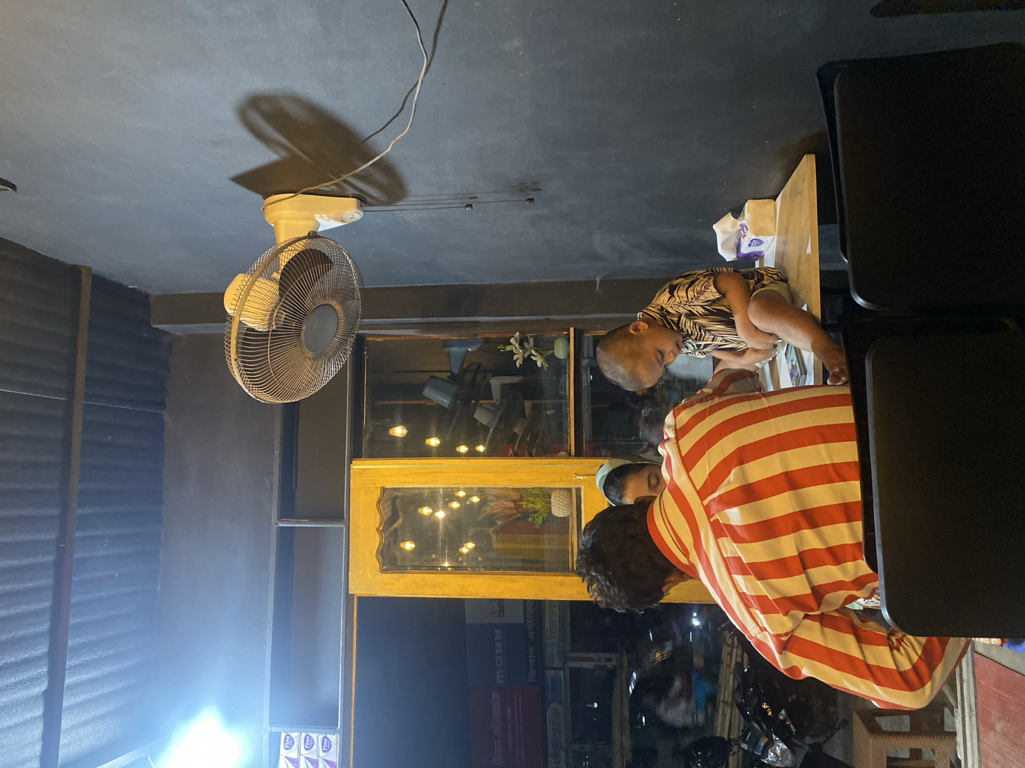 The image shows a scene inside a building. A child in a patterned shirt sits on a table, possibly eating. A person wearing a red and white striped shirt stands next to the table. There is a ceiling fan, and a mirror on the wall reflecting an exterior view. Outside the building, street signs are visible, one of which includes "MOHN" and the Bengali script "বিজ্ঞান জাদুকরী." There are also reflections of lights and vehicles in the mirror, suggesting a street scene, possibly in Dhaka, Bangladesh. There are chairs near the table.