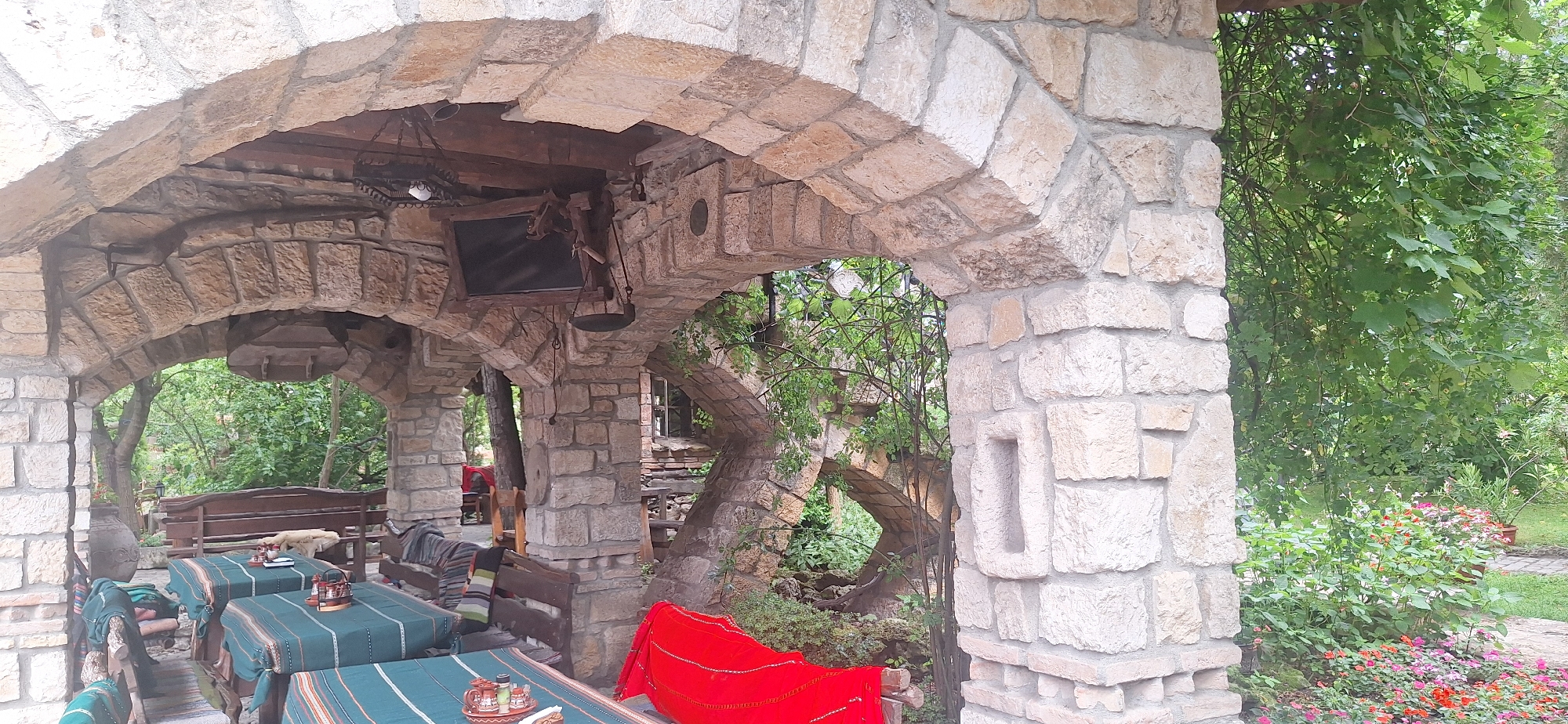 The image captures an outdoor seating area within what appears to be a restaurant or cafe, characterized by its rustic stone architecture. Located in the city of Kalimantsi, Bulgaria, the scene is set under a stone archway, with tables covered in green tablecloths and striped runners. Several chairs are arranged around the tables, some with blankets draped over them. In the background, lush green foliage and trees suggest a natural, perhaps garden-like, setting. A small, dark-colored television is mounted on the stone structure above one of the tables. The overall impression is one of a quaint, traditional outdoor dining space amidst nature.