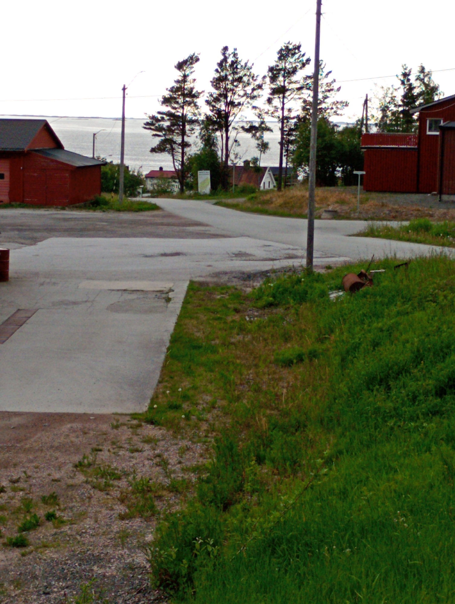 An outdoor, slightly overcast, daytime, medium-distance shot shows a rural landscape with several red buildings, trees, a body of water, and a paved area in the foreground. On the left, a long red building with a dark roof and a smaller attached red shed are visible. The main building has a window on its left side and a door on its right. In the middle ground, a light gray paved area extends from the foreground and curves to the right. To the right of the paved area, a taller red building with a dark roof and a white-framed window is visible. The background features a wide body of water under a bright sky, with trees lining the shore. The ground in the foreground is a mix of gravel and short green grass. A metal pole stands in the paved area. A brown, rusted metal barrel lies on its side in the grass to the right of the paved area, with some debris scattered around it.