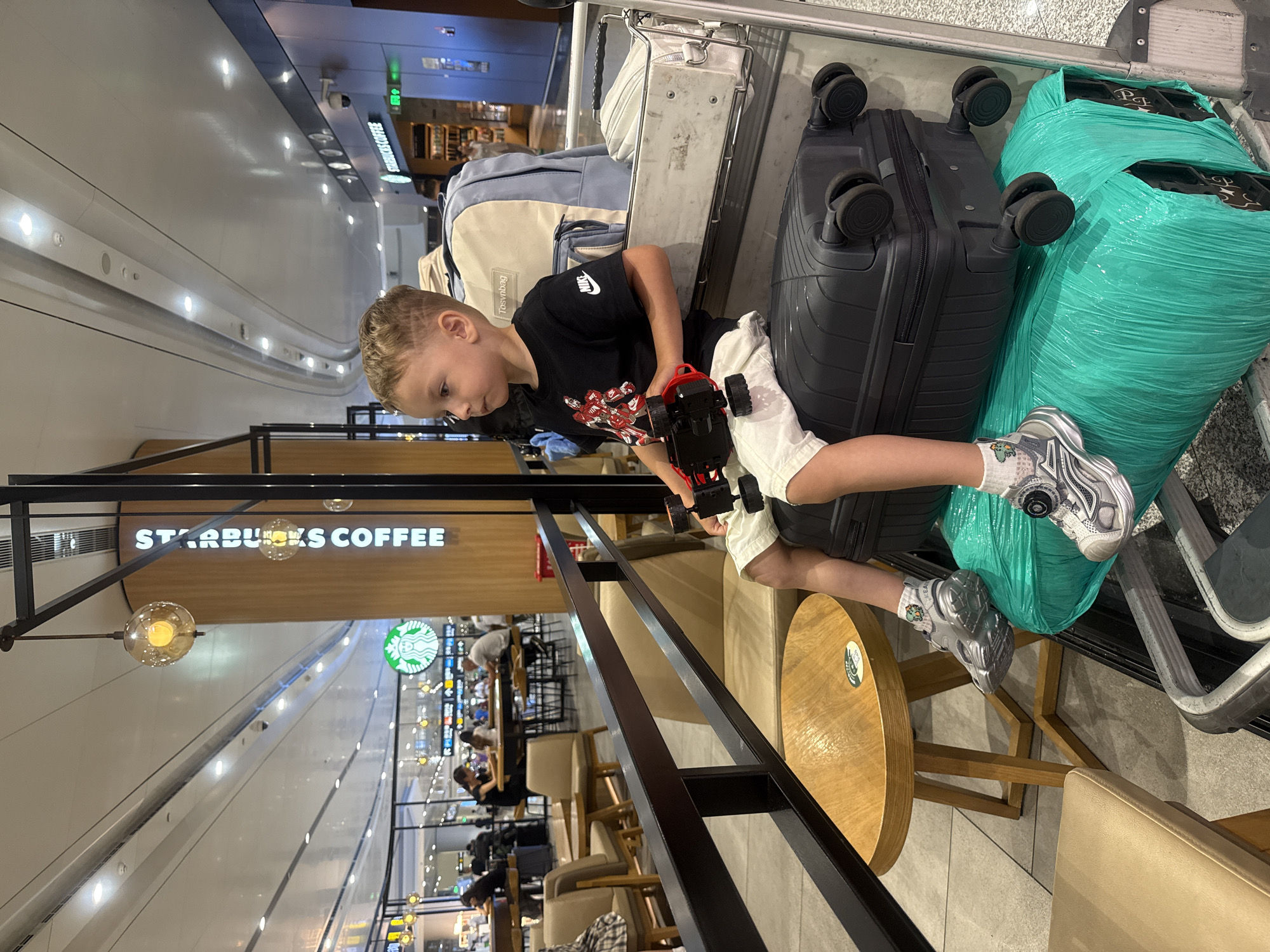 In Guangyang District, China, a young Caucasian boy with blonde hair, styled with a distinctive etched pattern on the side, is seated on a luggage cart in what appears to be an airport terminal. He is wearing a black t-shirt with a graphic design and white shorts, paired with silver and white sneakers. In his hands, he holds a red toy car.
The luggage cart is laden with several items, including a large black suitcase, a turquoise-wrapped package, and a lighter-colored backpack positioned behind him. To the left, a small round wooden table with a metallic base is visible, next to a tan upholstered chair.
In the background, the environment suggests a public indoor space, likely an airport, with a Starbucks coffee shop signage prominently displayed on a wooden pillar. The ceiling features modern lighting and ventilation systems. Other individuals and seating areas are visible in the distance, indicating a busy transit hub. The overall lighting is bright, typical of an airport terminal.