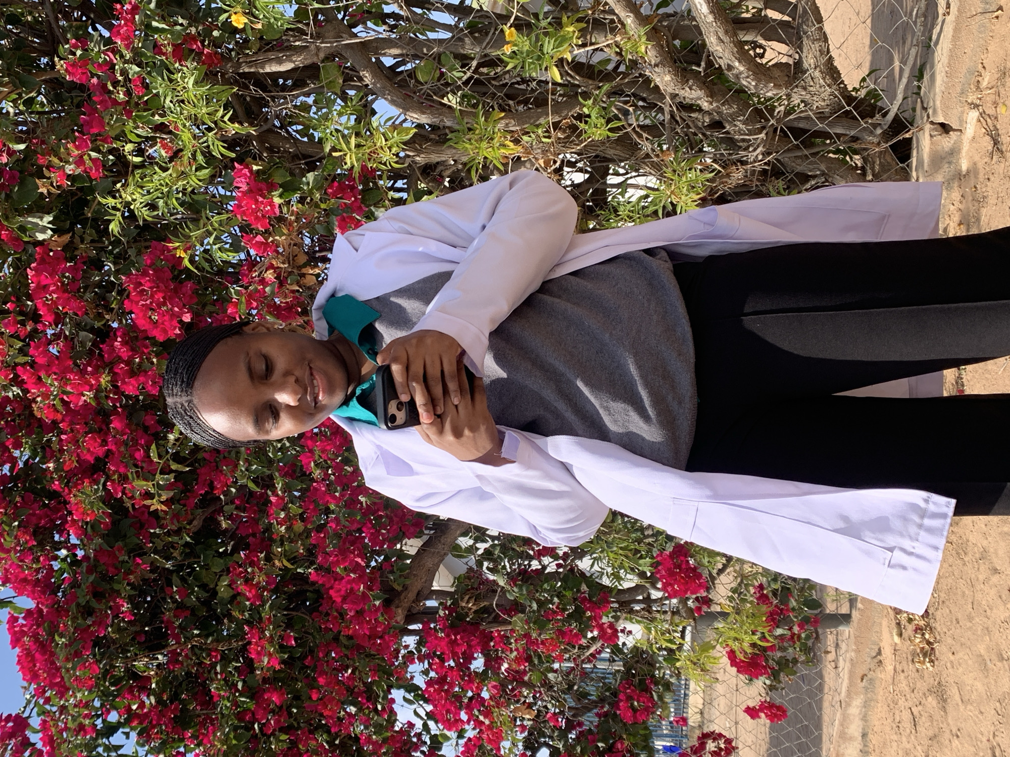 In Dodoma, Tanzania, a young Black female stands outdoors near a vibrant flowering tree, possibly a bougainvillea, with bright pink blossoms. She is wearing a white lab coat over a gray sweater and a teal collar. The woman is looking down at her black smartphone, which she holds in her hands. The ground is sandy with some dry leaves and sparse vegetation. A chain-link fence is visible behind her, and beyond that, a blue sky. The sun is shining, casting shadows and illuminating the scene.