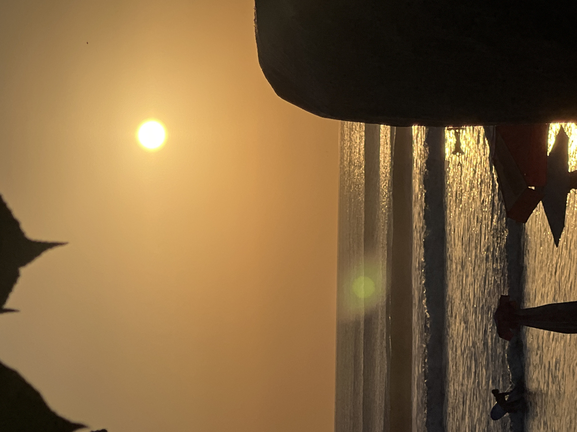 This is a photograph taken at Kuta Beach in Kuta, Indonesia, during sunset. The sun, a bright orb, is positioned in the upper right quadrant of the frame, casting a warm, golden glow across the scene. The sky is a hazy orange, with some dark, silhouetted shapes at the very top, possibly foliage. In the foreground, on the right, a large, dark, rounded object, likely a rock or a large piece of driftwood, is silhouetted against the sky.
The ocean occupies the middle ground, with multiple sets of waves rolling towards the shore. The water reflects the sunset, creating a shimmering effect. A notable lens flare, a green circular smudge, is visible over the ocean waves in the middle left of the image.
On the shore in the lower left, a person is visible, appearing to be a surfer or someone interacting with a surfboard. Their silhouette is dark. There are also some red objects in the sand, one of which appears to be a partially unfurled beach umbrella or shelter. The overall scene depicts a serene beach environment at dusk, with a focus on the natural beauty of the sunset over the ocean.