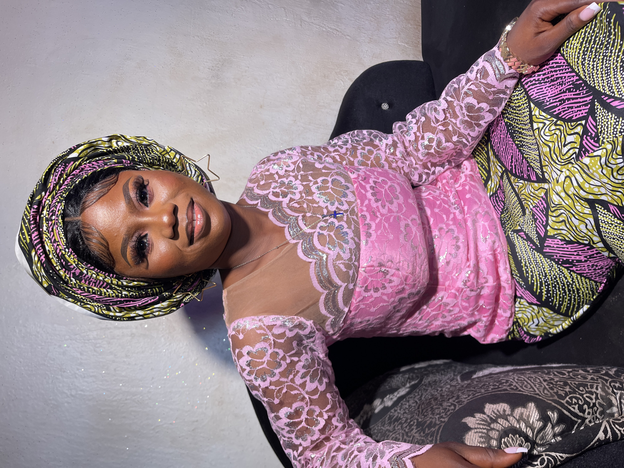 This image is a portrait of a smiling Black woman, likely captured indoors in Akwanga, Nigeria. She is seated, leaning back slightly in a dark upholstered chair or sofa, with her head gently tilted, engaging directly with the camera.
The woman is dressed in elegant attire, consistent with Nigerian traditional or celebratory fashion. She wears a vibrant headwrap (gele) with intricate patterns in black, yellow-green, and pink. Her top is a striking pink lace blouse, adorned with shimmering silver floral designs and featuring sheer nude mesh panels across the upper chest and shoulders, with long lace sleeves. Her lower garment, a wrapper (iro), matches the headwrap with a bold, abstract pattern in the same black, yellow-green, and pink colors.
Her accessories include a distinctive gold-toned, star-shaped wire earring on her left ear, a delicate silver necklace, and a gold-colored, braided-style bracelet on her right wrist. Her nails are manicured with classic French tips. She wears elaborate makeup, including defined eyebrows, detailed eye makeup with false lashes and pink eyeshadow, and glossy, light pink lips, contributing to her warm and confident expression.
The background features a light-colored, textured wall, possibly concrete or painted. Scattered reflective particles on the wall suggest either glitter or dust catching the light, adding a subtle sparkle. The lighting appears to be artificial, indicative of a professional or formal photo setting. The overall presentation suggests she is dressed for a special occasion or celebration. There is no visible text in the image.