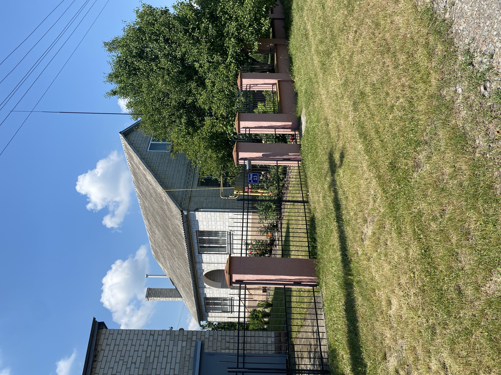 In Vasylky, Ukraine, a light brick house with a gray, corrugated roof is visible behind a black metal fence. The house has multiple windows with bars, a prominent brick chimney, and an arched entranceway. In front of the house, there are rose bushes and other low-lying plants. A street sign with the inscription "вул. Леніна 23" is attached to one of the pink-topped fence posts. To the left of the house, another building, partially visible, is constructed from lighter colored bricks. A large green tree stands to the right of the house, partially obscuring the view of the sky, which is blue with scattered white clouds. Power lines are strung across the upper right portion of the image. The foreground is a grassy lawn with patches of dry grass.
