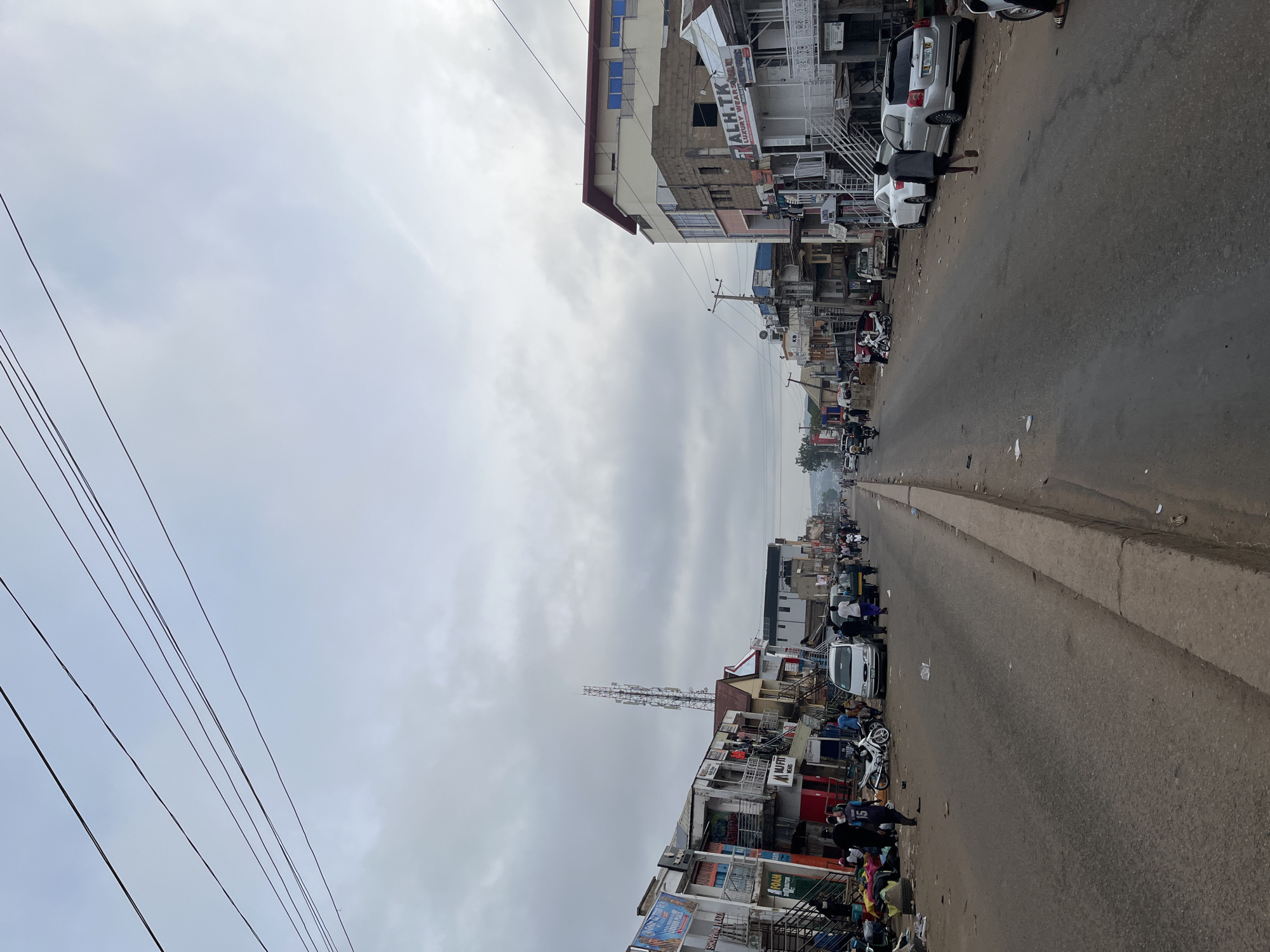 The image depicts a street in Jos, Nigeria. The road stretches into the distance, lined on both sides by multi-story buildings. Several vehicles, including cars and motorcycles, occupy the street. A cellular tower rises above the buildings on the left. Overhead electrical wires crisscross the sky. The sky is overcast with grey clouds. Pedestrians walk along the roadside. Litter is visible on the road and sidewalks. Buildings display signage.