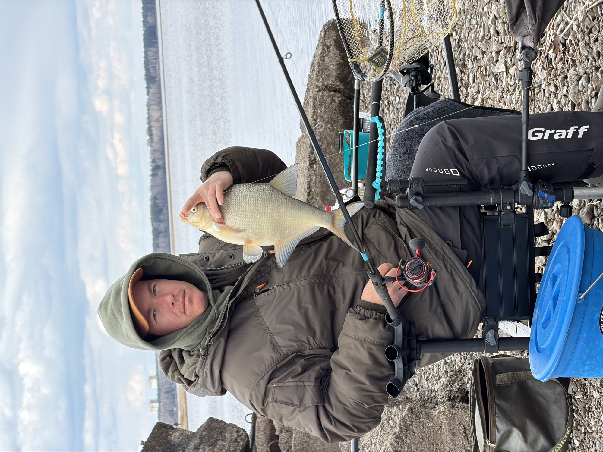 A man is seated by a large body of water, proudly holding a fish he has caught. The image is taken outdoors on what appears to be a cool, overcast day.
The man, likely a fisherman, is in the foreground, facing slightly towards the camera with a content expression. He wears a dark green hooded jacket with a brown cap underneath the hood. His right hand supports the belly of a light-colored fish, which appears to be a bream or similar carp-like species, showcasing its prominent scales and fins. The fish is held horizontally, occupying a significant part of the frame.
He is positioned on a specialized fishing chair or platform, equipped with various accessories. A black fishing rod with blue details is visible, extending diagonally across the frame, with a black and red fishing reel attached. Nearby, a yellow-meshed landing net with a tag reading "ULTRA LITE" is hanging. A sturdy black fabric cover or bag, with the brand name "Graff" prominently displayed in white, forms part of his setup. To his right, a bright blue bucket is placed on the ground, and a green bag with "AQUATIC" visible on its side lies next to it. Several rod holders are integrated into his fishing station. The man's jacket also has a small tag on the zipper with the text "URSA".
The background reveals a wide expanse of water, presumably a river or lake given the location context of Chaikovsky, Russia. The shoreline consists of small rocks or pebbles. On the distant bank, a faint treeline can be observed, separating the water from the cloudy, light blue sky. The overall scene depicts a successful fishing outing.