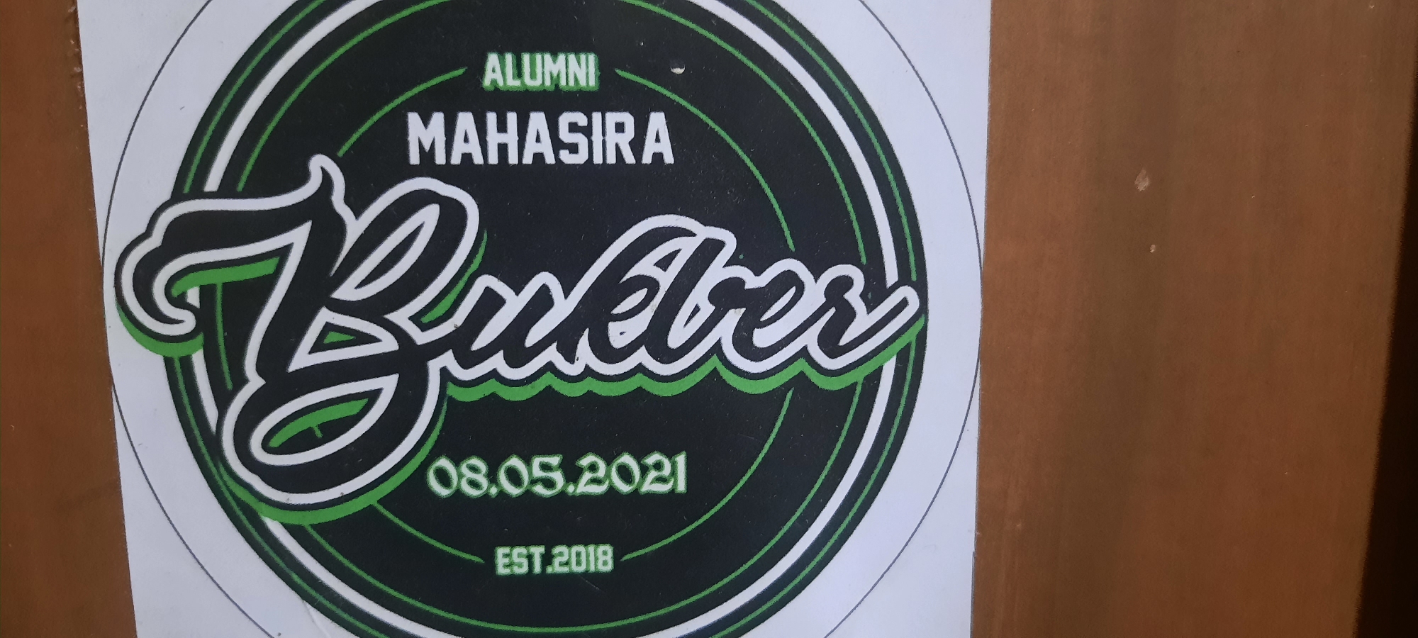 The image is a close-up of a circular sticker with a black background and green and white accents. The sticker has a logo that appears to be for an alumni association called "ALUMNI MAHASIRA". Below this text, a large, stylized cursive word "Bukber" is prominently displayed in white with a green outline. Beneath "Bukber", the date "08.05.2021" is written in green. At the bottom of the sticker, "EST.2018" is also in green. The sticker is affixed to a wooden surface, likely a door or wall, which is brown and has some light-colored specks. The sticker is positioned on the left side of the frame, with the wooden surface occupying the right side. The lighting suggests an indoor environment. No people are visible in the image, nor is there any specific indication of the city of Ploso, Indonesia within the visual content of the sticker itself.