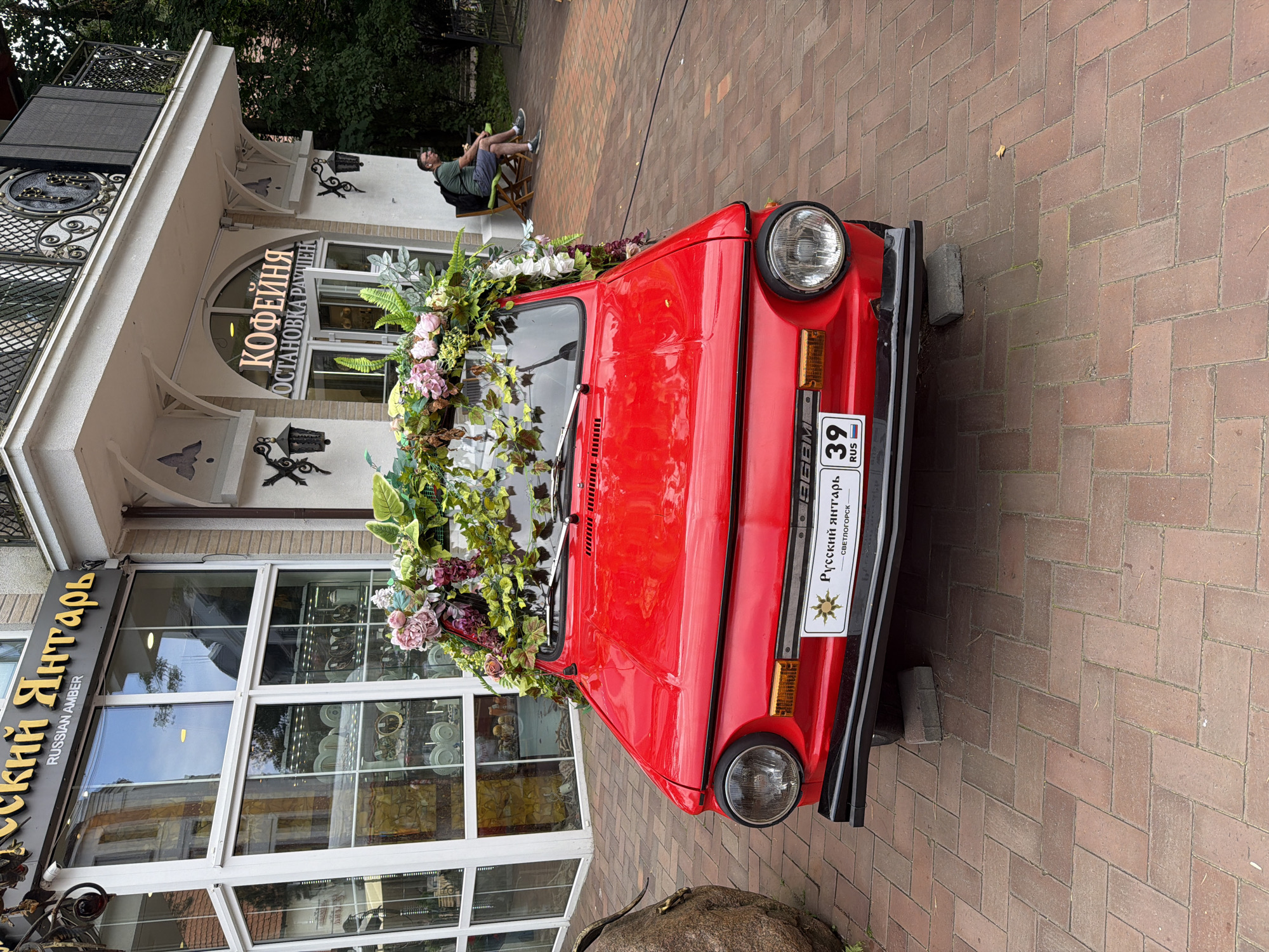 A red ZAZ 968M car, decorated with a floral arrangement, is parked on a brick-paved sidewalk in front of a building in Svetlogorsk, Russia. The building has signage that reads "РУССКИЙ ЯНТАРЬ" (Russian Amber) and "КОФЕЙНЯ" (Coffee Shop). A man is seated on a chair in the background to the right, near the entrance to the coffee shop. The car's license plate is visible and reads "39 RUS." The overall scene appears to be a commercial area with a vintage car serving as a decorative element.