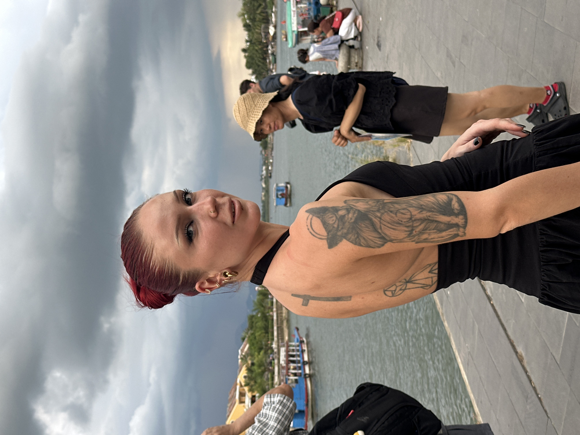 The image shows a woman with red hair styled in a bun, facing away from the camera but looking over her shoulder. She has multiple piercings and tattoos on her upper back and arm. She is wearing a black dress with ruffled hem. In the background, there is a body of water with boats, and buildings with yellow facades are visible on the opposite bank. Other people are seen in the background, including a woman wearing a straw hat and a black top and skirt. The scene appears to be along the Thu Bon River in Hoi An, Vietnam, a city known for its historic architecture and vibrant riverfront. The sky is cloudy, suggesting overcast weather.