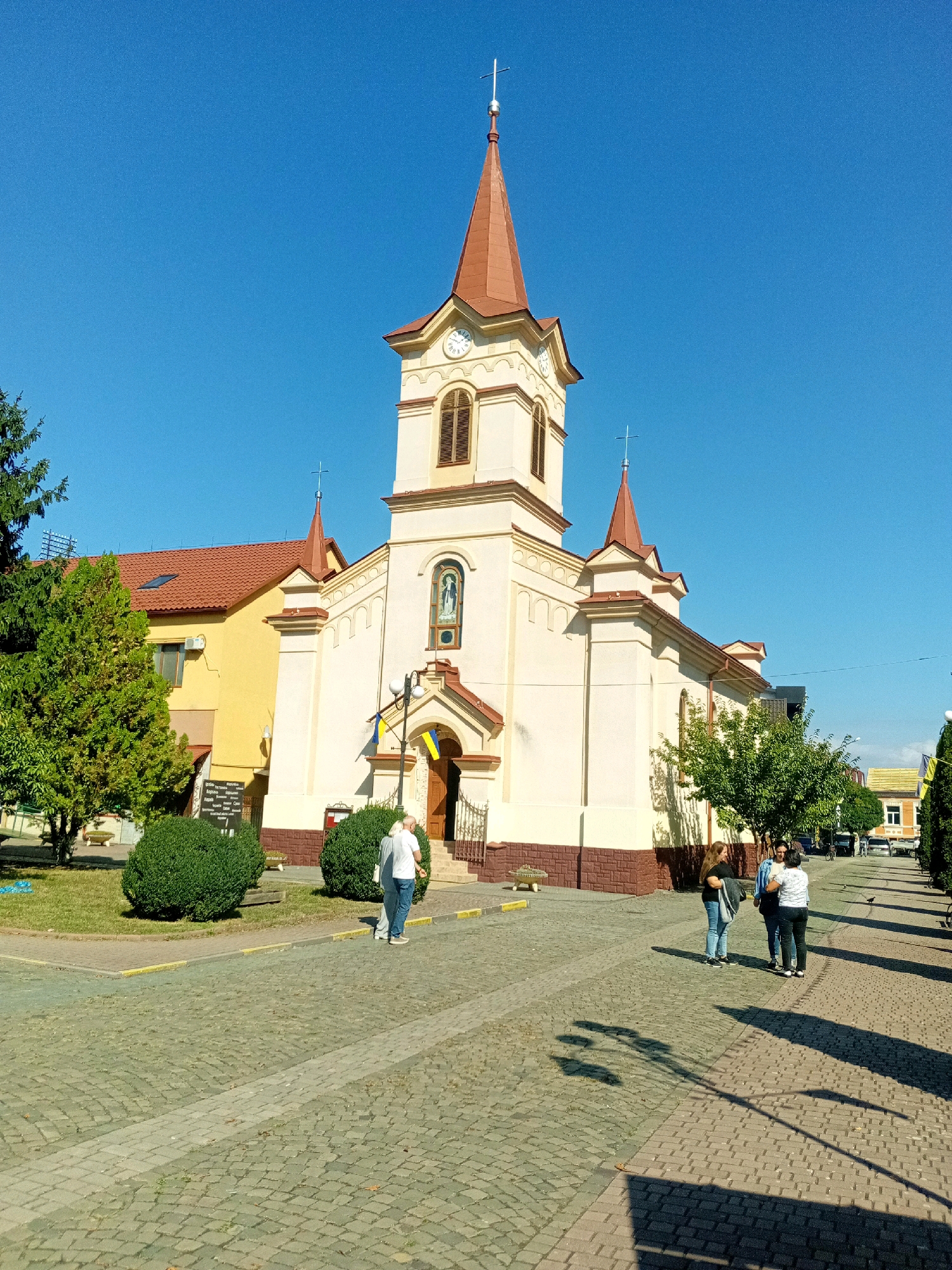 The image captures a sunny day in Tiachiv, Ukraine, focusing on a distinctive church with a tall, pointed red roof and a clock tower. The church is painted cream with brown trim and features a small statue of a religious figure above its entrance, which is flanked by two Ukrainian flags. The foreground shows a cobblestone walkway with two people walking away from the camera and three people standing and talking. To the left of the church, a bright yellow building with a red roof is visible, and several well-maintained green bushes are planted in front of it. Trees with green foliage are scattered throughout the scene, casting shadows on the walkway. The sky is a clear, vibrant blue.
