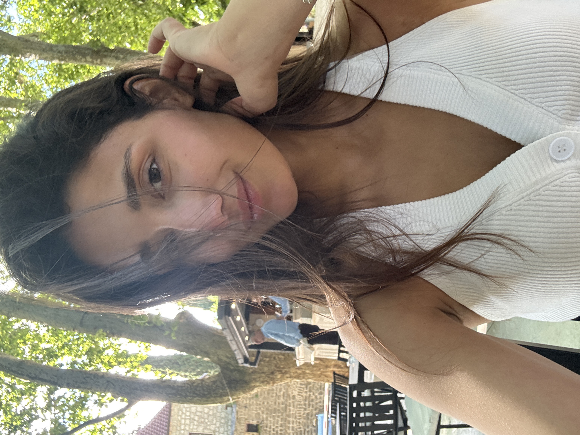 The image shows a close-up, selfie-style shot of a young woman with long, dark hair and fair skin, wearing a white ribbed tank top. She is outdoors, with dappled sunlight filtering through green trees. Her right hand is raised, touching her ear and hair. In the blurred background, a man in a light blue shirt and dark pants stands near a structure, possibly a food stall or kiosk. Behind him is a stone building with a red-tiled roof. Several outdoor tables and chairs are visible in the foreground. The setting appears to be a cafe or restaurant with an outdoor seating area, likely in Trebinje, Bosnia and Herzegovina, given the presence of sycamore trees which are characteristic of the region and the architecture of the building in the background.