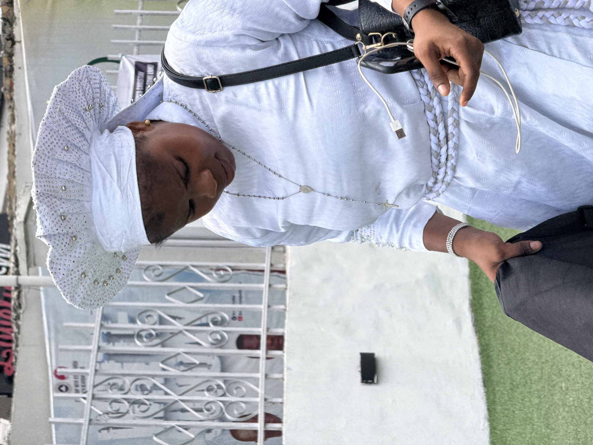 A medium-shot depicts a person in Ado, Nigeria, wearing a traditional white attire with a sequined head covering. The attire has a braided white and gray pattern down the center. A black purse with a strap is slung over the shoulder, and a phone with a charging cable is visible near the person's hand. The person wears a silver rosary necklace and a beaded bracelet. The backdrop includes a white building with decorative iron bars and green artificial turf.