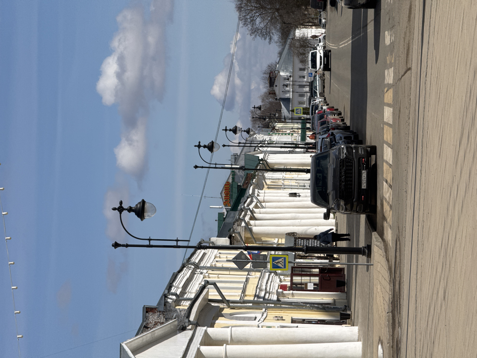 The image shows a street in a city. A line of parked cars is along the right side of the street. A crosswalk is visible. On the left, a building with white columns lines the street, with black lamp posts in front of the building. A person is standing on the sidewalk. The sky is blue with some clouds. The location is Unknown, Unknown.