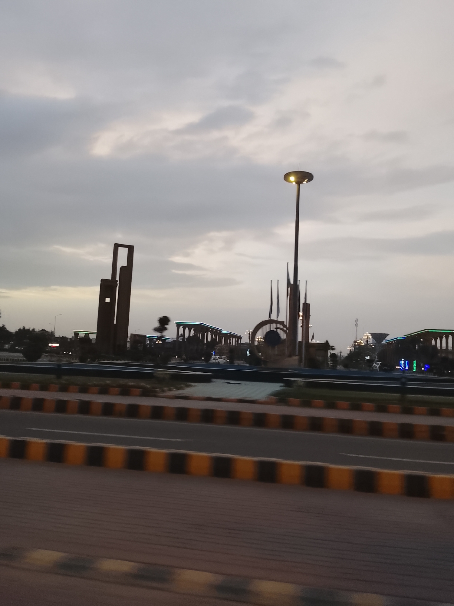 This media file captures a modern cityscape at dusk in Al Falah Modern City, Pakistan. The scene appears to be a large intersection or roundabout, viewed from a moving vehicle, indicated by slight motion blur in the foreground.
The sky is overcast, with soft, diffused light, and faint hints of lighter clouds. The artificial lights of the city have begun to illuminate, suggesting it's either late evening or early morning.
In the center of the image, on a wide median or central island, are several prominent architectural and sculptural elements. To the left, a tall, dark, abstract monument with a rectangular opening stands against the sky. Further to the right, a striking, tall streetlamp with a circular, glowing yellow light fixture dominates the central area. Behind it, a large, dark circular archway is visible, adorned with what appear to be flags or banners. Other buildings and structures in the background feature archways and are lit with decorative green and blue lights, contributing to the modern aesthetic of the city. Trees and shrubs are planted on the median, adding greenery to the urban landscape.
In the immediate foreground, a road with distinct yellow and black striped curbs or barriers runs across the lower part of the frame, alongside a paved sidewalk area. No people or visible text are discernible in the image. The overall impression is one of a well-developed, contemporary urban environment.