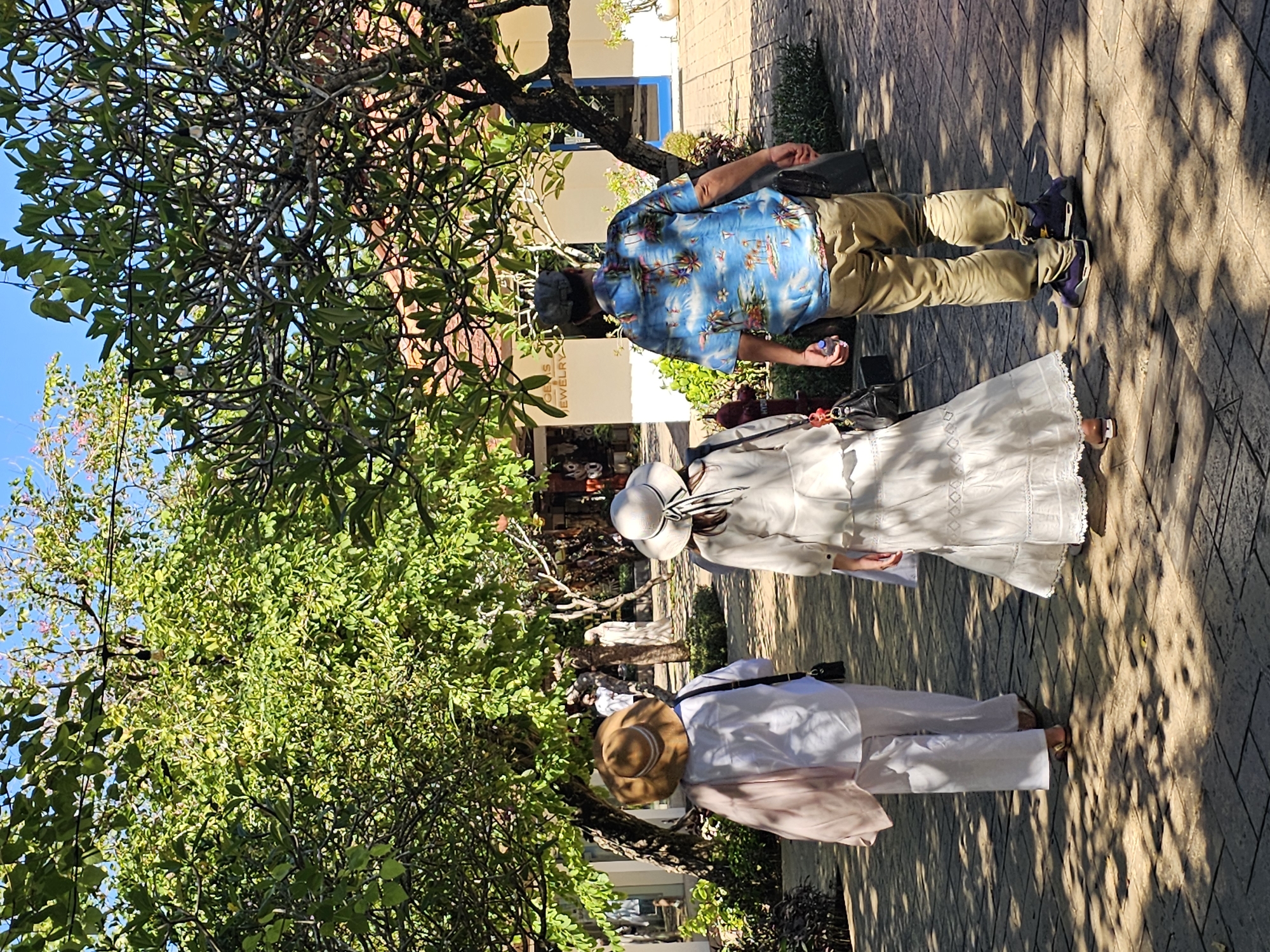The image depicts three people walking outdoors on a paved path. The first person, closer to the camera, wears a wide-brimmed straw hat, a white long-sleeved shirt, white pants, and sandals. A light pink garment or shawl is draped over their shoulder. The second person, in the middle, wears a white dress with embroidered details, a white hat with a black ribbon, and brown sandals. The third person, furthest from the camera, is a man wearing a blue Hawaiian shirt, khaki pants, and purple sneakers. The setting appears to be a commercial area with a jewelry store visible in the background. The scene is heavily shaded by trees. Considering the presence of "Jewelry" signage, and the overall style of architecture, it's plausible that the location is somewhere in the tourist-oriented areas of Kuta Selatan, Indonesia.