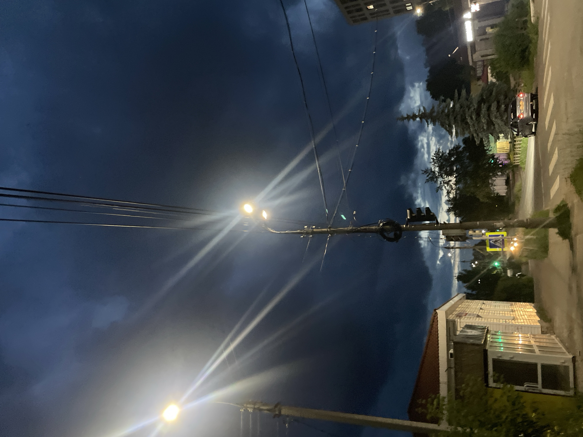The image shows a street scene at dusk or night in Votkinsk, Russia. The sky is dark blue with some clouds, illuminated by streetlights. A tall, thin evergreen tree stands prominently in the mid-ground. To the right of the tree is a paved road with a pedestrian crossing marked by white stripes. A dark-colored car with its taillights on is parked on the side of the road. In the foreground and along the right edge of the image, a building with a red roof and white walls is visible, featuring large windows that reflect the light. Several utility poles with wires are present, with streetlights attached to them. One streetlight is particularly bright, creating lens flares. The overall impression is a quiet, urban evening. There is no visible text in the image.