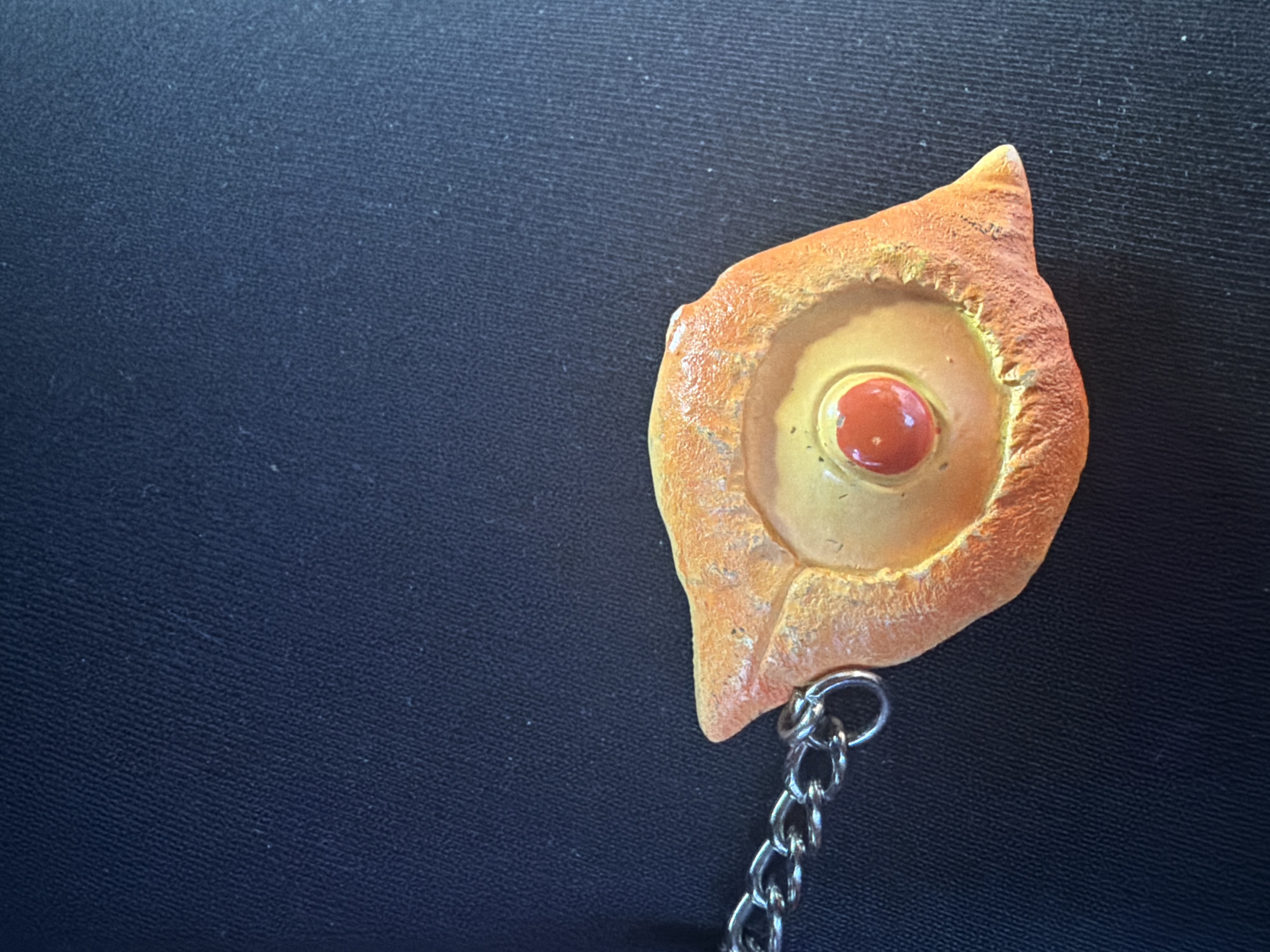The image shows a close-up of an orange, eye-shaped pendant with a red bead in the center, attached to a silver chain. The pendant has a textured, slightly rough surface. The background is a dark, solid fabric, possibly clothing. There is no discernible indication of the city of Yerevan, Armenia in the image.