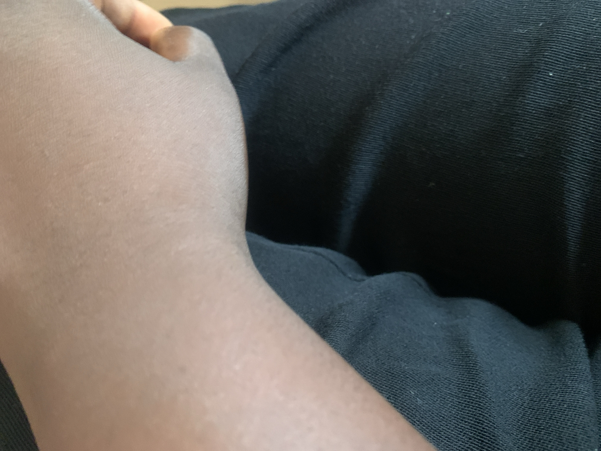 The image shows a close-up view of a person's forearm and hand resting on dark fabric, likely clothing. The skin tone is medium to dark brown. The texture of the skin is visible, with some slight pores or irregularities. The hand is partially visible on the right side of the frame, with the thumb and part of the index finger extended. The dark fabric appears to be woven, with a ribbed texture visible in some areas. The lighting suggests an indoor setting, with soft illumination casting subtle shadows. The image is focused on the arm and fabric, with no background details visible. There are no discernible elements that would indicate a specific location within Jauro Yinu, Nigeria.