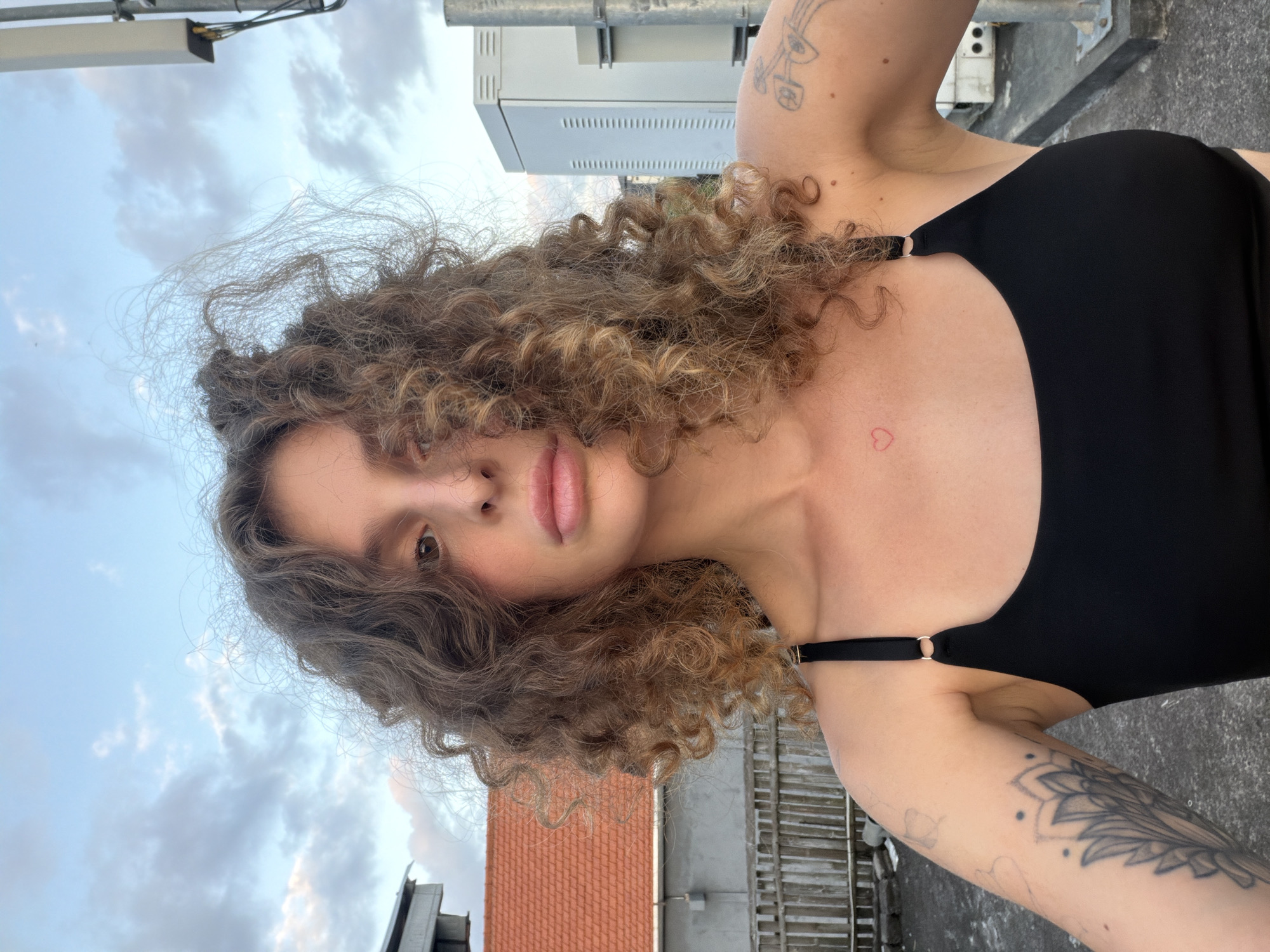 An outdoor, eye-level, close-up, selfie portrait shows a young Caucasian woman with voluminous, curly, brown hair and a light tan complexion. She is wearing a black, sleeveless top. A small, red heart tattoo is visible on her upper chest, and she has tattoos on her left arm. She is looking directly at the camera with parted lips. Her body is angled slightly to the left, and her head is tilted forward. The background shows a cloudy sky with a glimpse of an orange-tiled roof and a gray building to the left. To the right, there is a tall, gray utility pole and a white electrical box. The scene appears to be on a rooftop in the city of North Kuta, Indonesia.