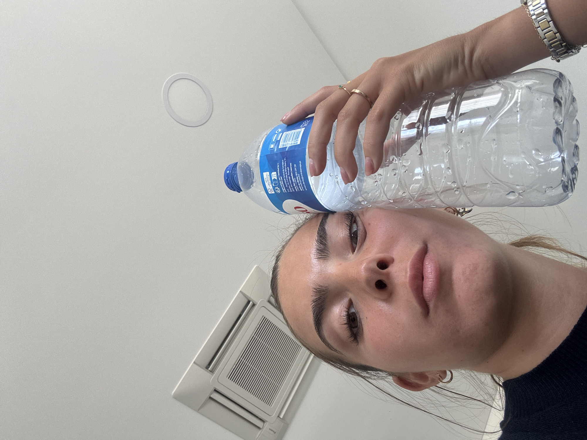 A low-angle shot features a woman holding a plastic water bottle against her forehead. Her hand, adorned with rings and a metal watch, secures the bottle. The background is a white ceiling with a circular light fixture and a square air conditioning vent. Text on the bottle is readable, suggesting the water is bottled by Sociedade Central de Cervejas e SCCA in Estrada do Alfarrobeiro, Portugal.