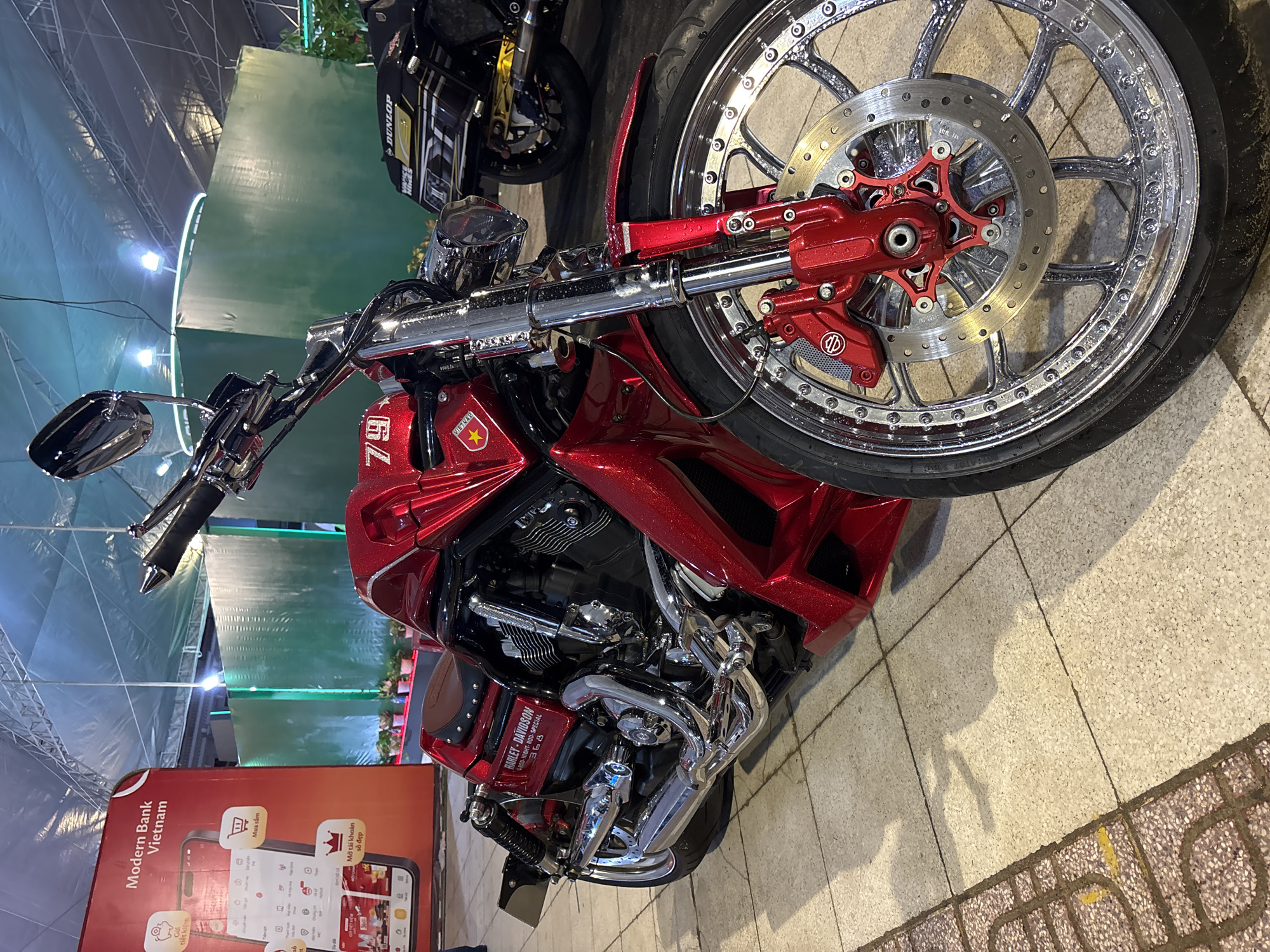 The image shows a red Harley-Davidson V-Rod Night Rod Special motorcycle on a tiled surface. The front wheel is prominently displayed, with a complex chrome rim and red brake calipers. A second motorcycle is partially visible behind it. A sign for "Modern Bank Vietnam" is in the foreground. A sign reads "Nha Trang Ward, Vietnam." The environment suggests an indoor display or showroom setting, with additional lighting and tented walls visible.
