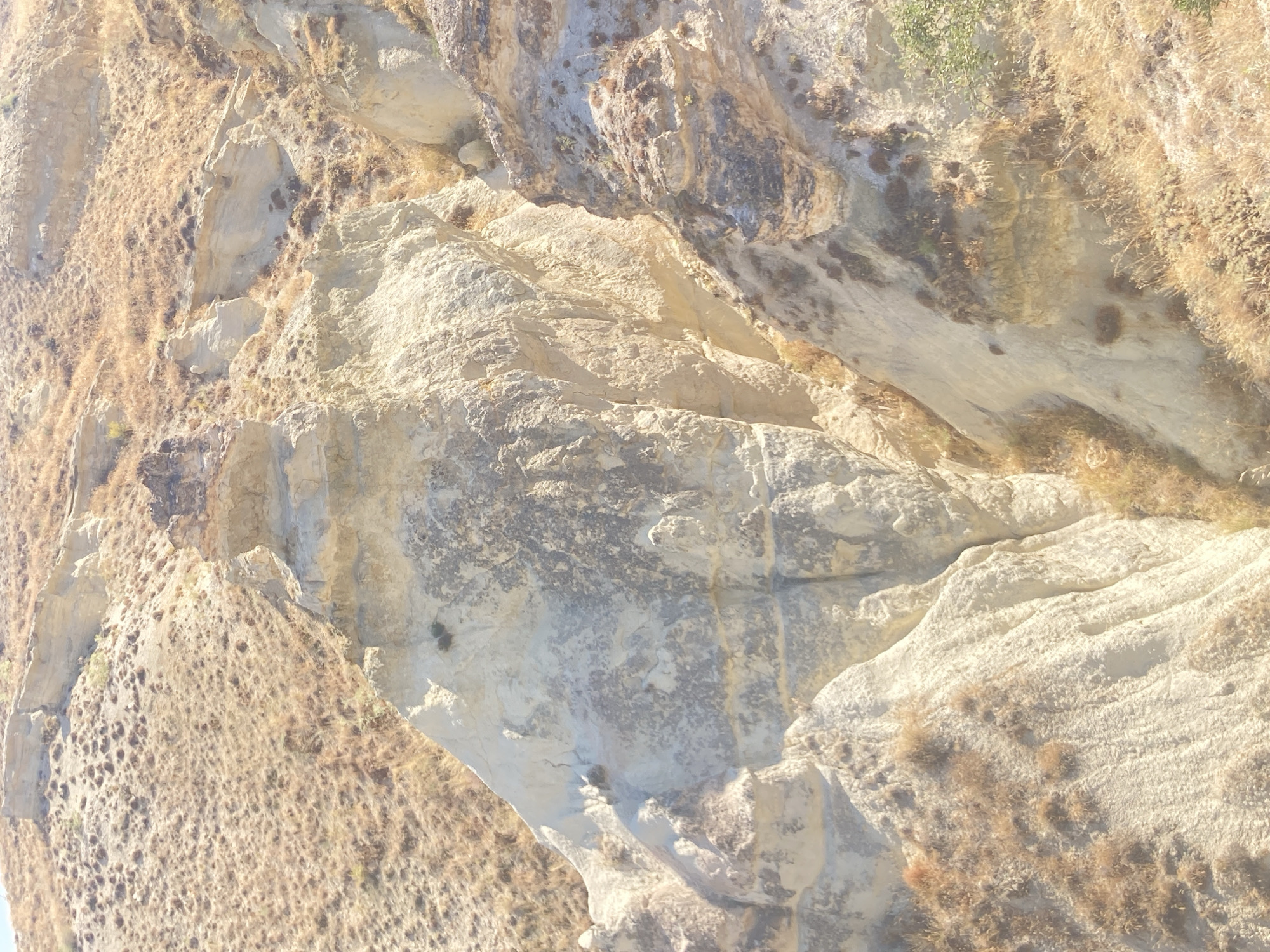 The image displays a geological formation in Ürgüp, Turkey, characterized by its undulating terrain and distinct rock structures. The foreground is dominated by a tall, weathered rock pillar, intricately carved by natural elements, showcasing layered strata of light-colored stone. Surrounding this central formation are steep, eroded slopes covered in dry, golden-brown vegetation, typical of the region's arid climate. The landscape suggests a dry, sunny day with ample sunlight casting shadows that accentuate the rock's texture and depth. The overall impression is one of a rugged, natural environment, possibly a part of Cappadocia's iconic fairy chimney landscape.